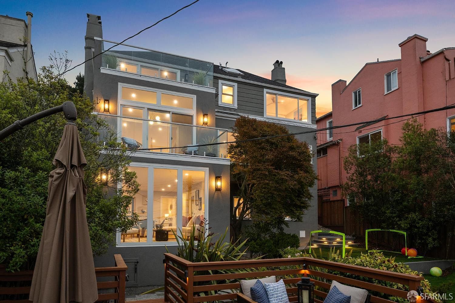 This is a rear view of a multi-story home, showcasing a modern design with large windows and a glass-railed balcony. The exterior is painted in a neutral gray tone, complemented by a well-maintained backyard featuring a wooden deck, lush greenery, and outdoor seating. The scene is set during twilight, with warm interior lights creating an inviting ambiance.