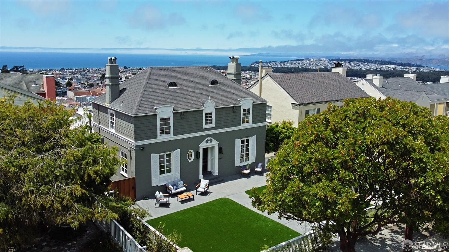 This is an exterior front view of a two-story house with a gray facade and white trim. The house features a well-manicured lawn, a patio area with outdoor seating, and a view of the city and ocean in the background. The architectural style is traditional with dormer windows and a symmetrical design, creating a sophisticated and inviting impression.