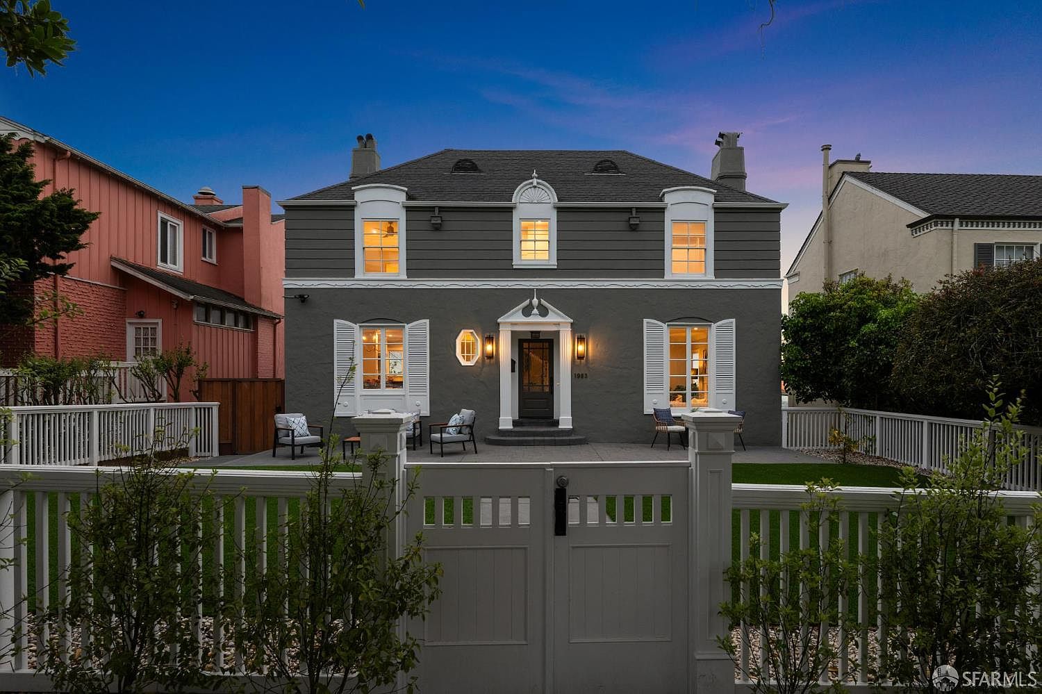The image showcases the front exterior of a two-story house with a gray facade and white trim. The house features symmetrical design elements, including windows with white shutters and a central front door with decorative lighting. A white picket fence encloses the front yard, adding to the property's curb appeal and creating a welcoming atmosphere.