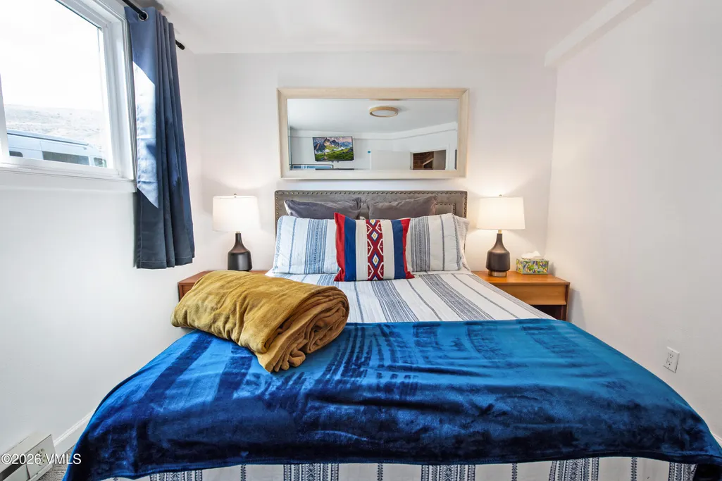 This cozy bedroom features a full bed dressed with blue-and-white striped bedding and an eye-catching, colorful decorative pillow. The room is symmetrically arranged with a matching pair of modern bedside tables and lamps, and includes a large mirror above the bed that brightens the space. A plush gold throw blanket adds a warm accent to the blue velvet-style coverlet, creating a comfortable and inviting atmosphere.