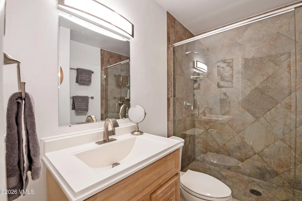This functional bathroom features a light wood vanity topped with a clean, white integrated sink and a modern brushed-nickel faucet. A walk-in shower with large, diagonal-set earth-toned tile adds a rustic, textured element, while the frameless glass enclosure keeps the space feeling open and bright. A magnifying mirror on the counter and well-placed lighting fixtures complete the practical and tidy design.