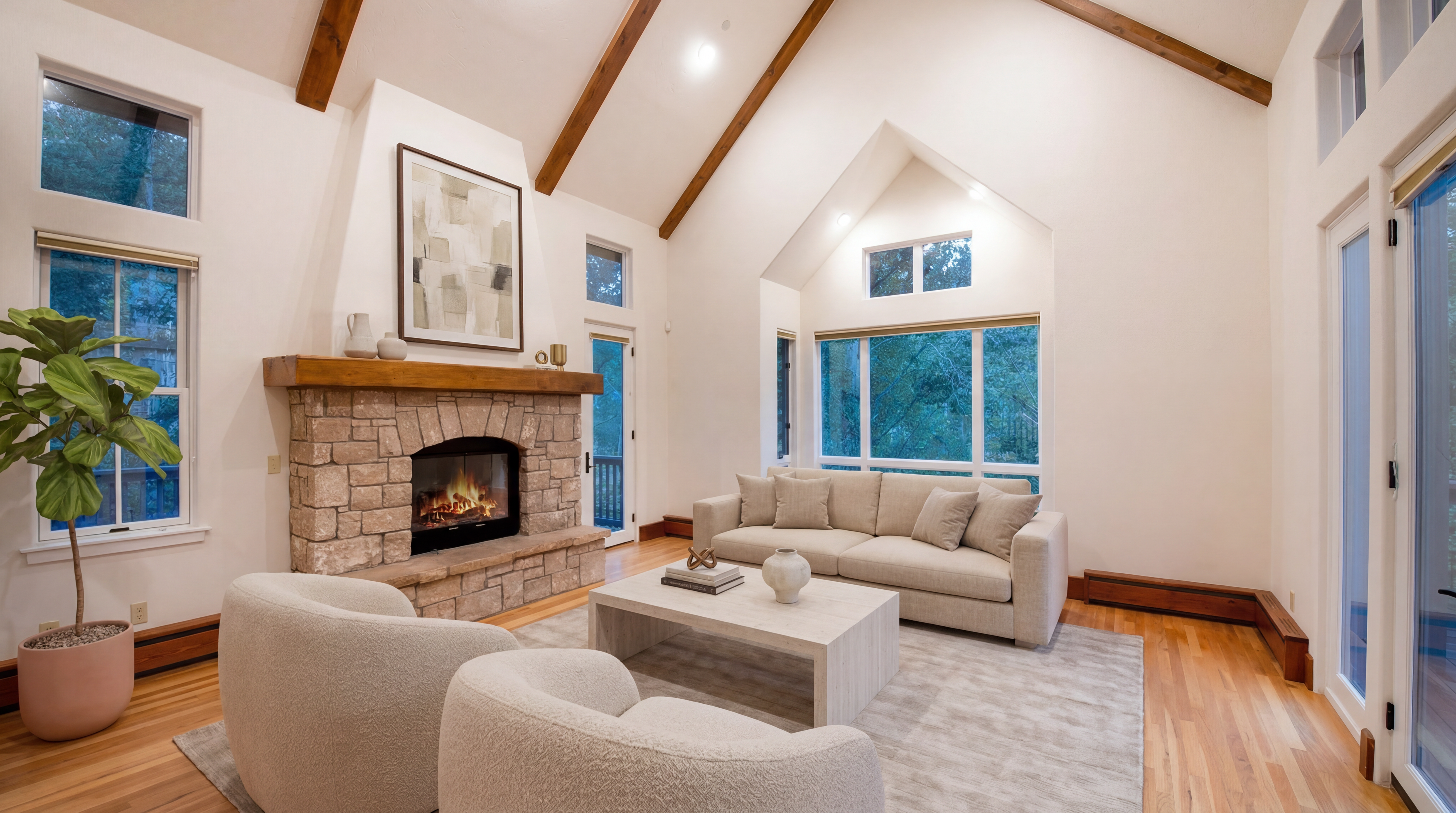 This living room features a striking vaulted ceiling with exposed wooden beams and a warm stone fireplace as its centerpiece. A neutral color palette, plush modern furniture, and large windows create an airy, comfortable, and inviting atmosphere. The space seamlessly blends architectural character with contemporary design, perfect for relaxation or entertaining.