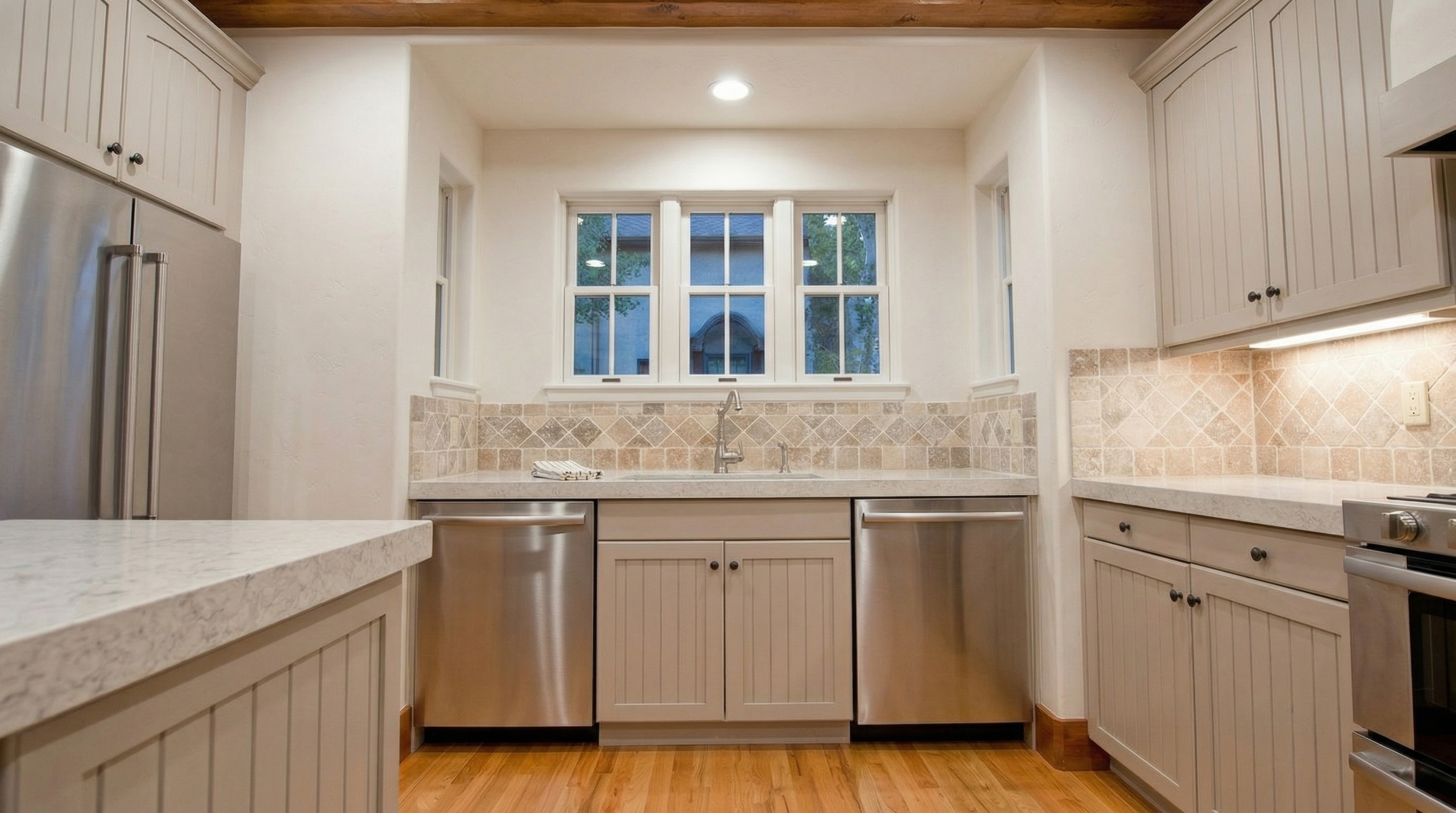 This kitchen features a warm, traditional aesthetic with light-toned, beadboard-style cabinetry and neutral travertine-patterned stone backsplash. The space is equipped with modern stainless steel appliances, including a double-dishwasher setup flanking the central sink, and boasts elegant stone countertops atop a polished hardwood floor. Exposed wooden beams on the ceiling add a touch of rustic charm, creating a cohesive and inviting culinary environment.
