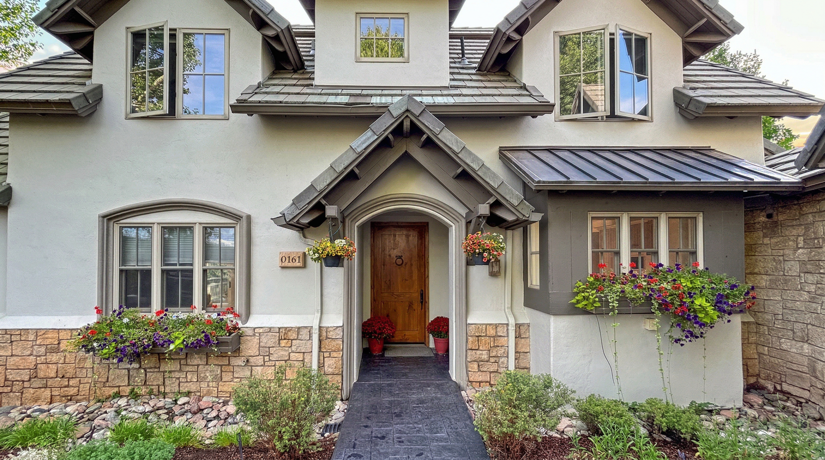 This charming front exterior features a blend of stucco and rustic stonework, giving it a classic, elegant cottage-inspired aesthetic. The home boasts a prominent wooden front door sheltered by a decorative portico, flanked by vibrant window boxes filled with colorful flowers and lush landscaping. The symmetrical roofline, detailed masonry, and well-maintained entry path create an inviting and sophisticated curb appeal.