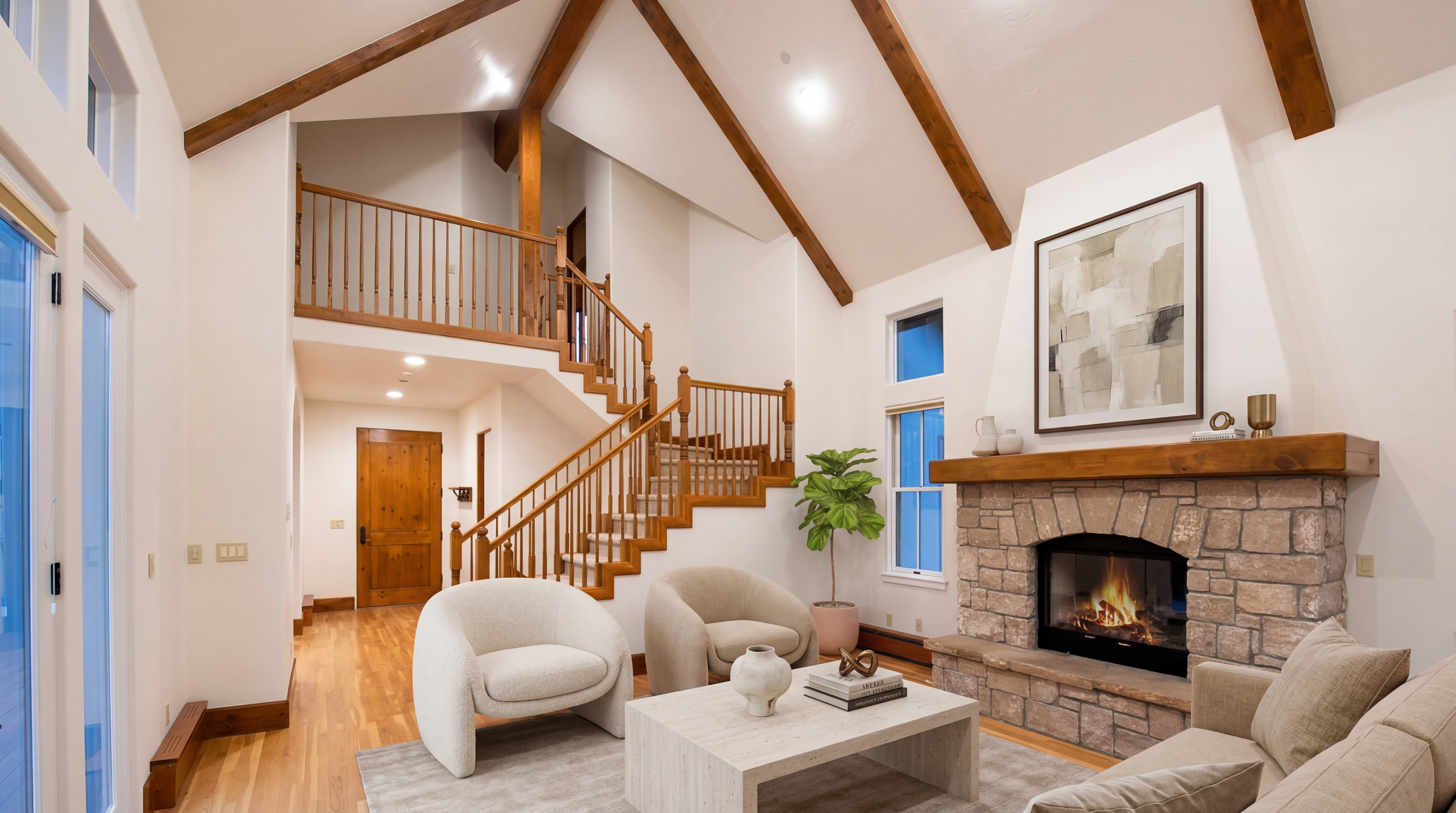This bright and inviting living room features a striking stone fireplace with a rustic wood mantel, serving as a focal point beneath a soaring, vaulted ceiling with exposed wood beams. The open-concept space flows seamlessly into a staircase with classic wood railings and includes comfortable modern seating arranged around a minimalist coffee table. Natural light floods the room through expansive windows, creating an airy, open feel that highlights the warm wood and stone textures throughout.