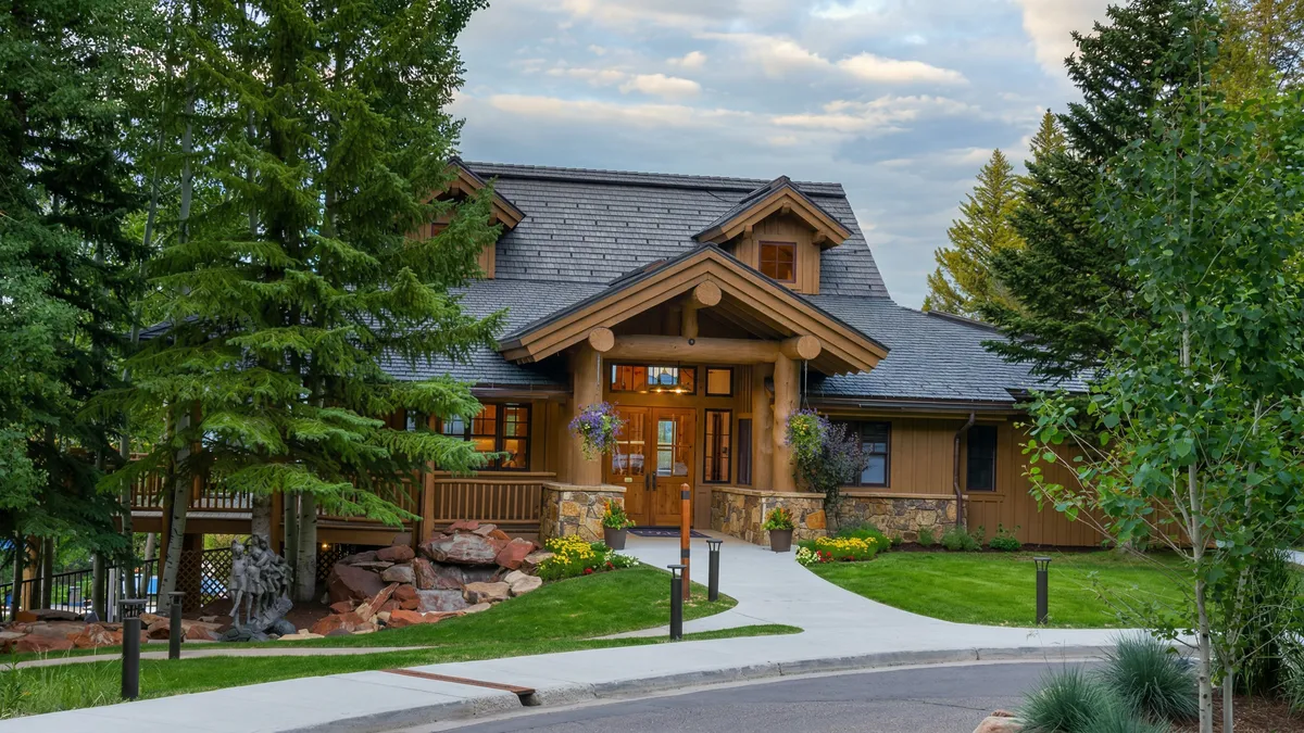 This charming lodge-style home features a rustic exterior with log accents, stone masonry, and a dark shingled multi-gabled roof. The front entrance is welcoming, framed by substantial log beams and complemented by a manicured walkway, vibrant landscaping, and lush surrounding trees. The overall impression is a classic, upscale mountain aesthetic perfect for a high-end retreat.