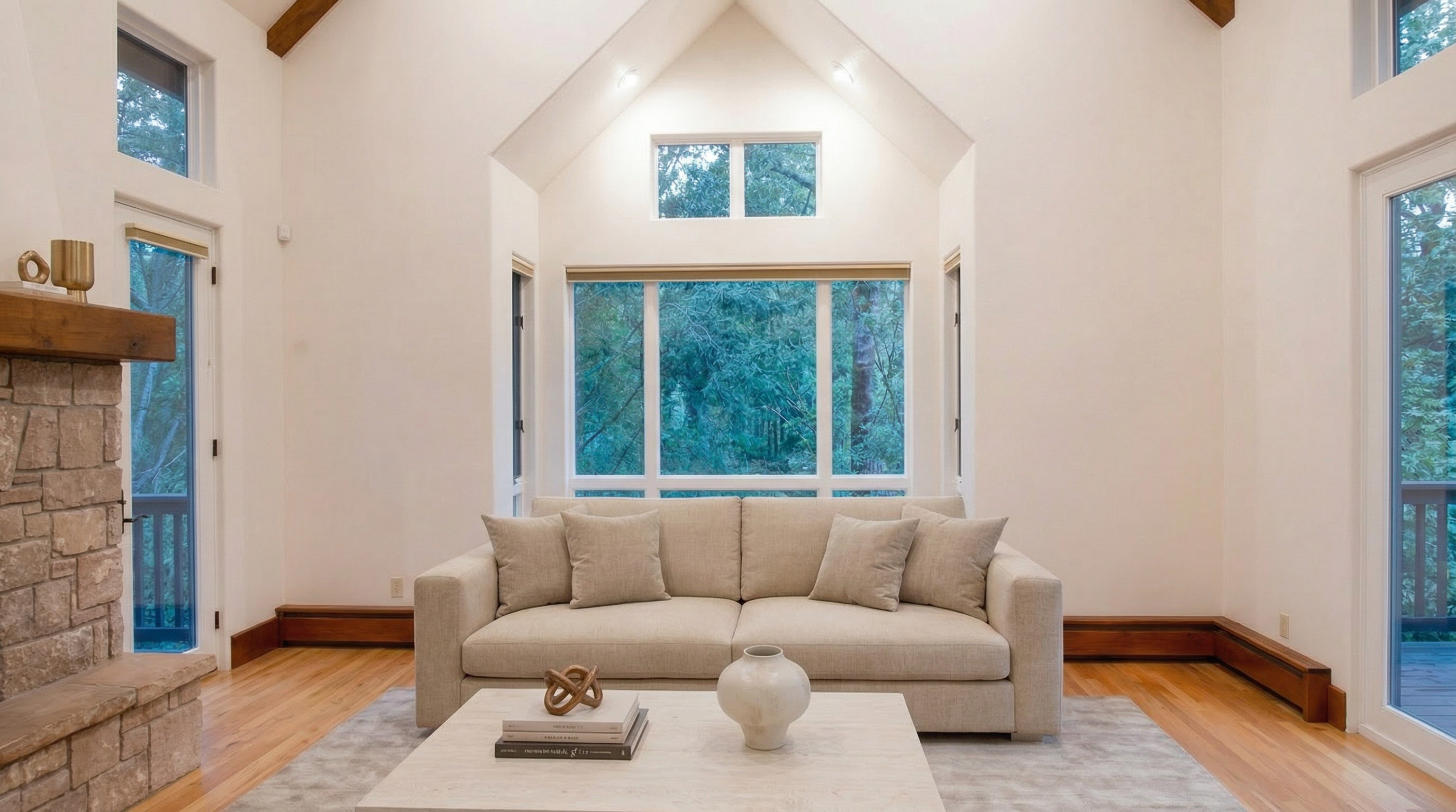 This bright and airy living room features high vaulted ceilings, a large neutral sofa, and a stone-faced fireplace with a wood mantel. Large windows frame a beautiful view of lush green trees, creating a serene and natural atmosphere. The room is accented with minimalist decor, including a sleek coffee table with stacked books and a ceramic vase, providing a clean, contemporary aesthetic.
