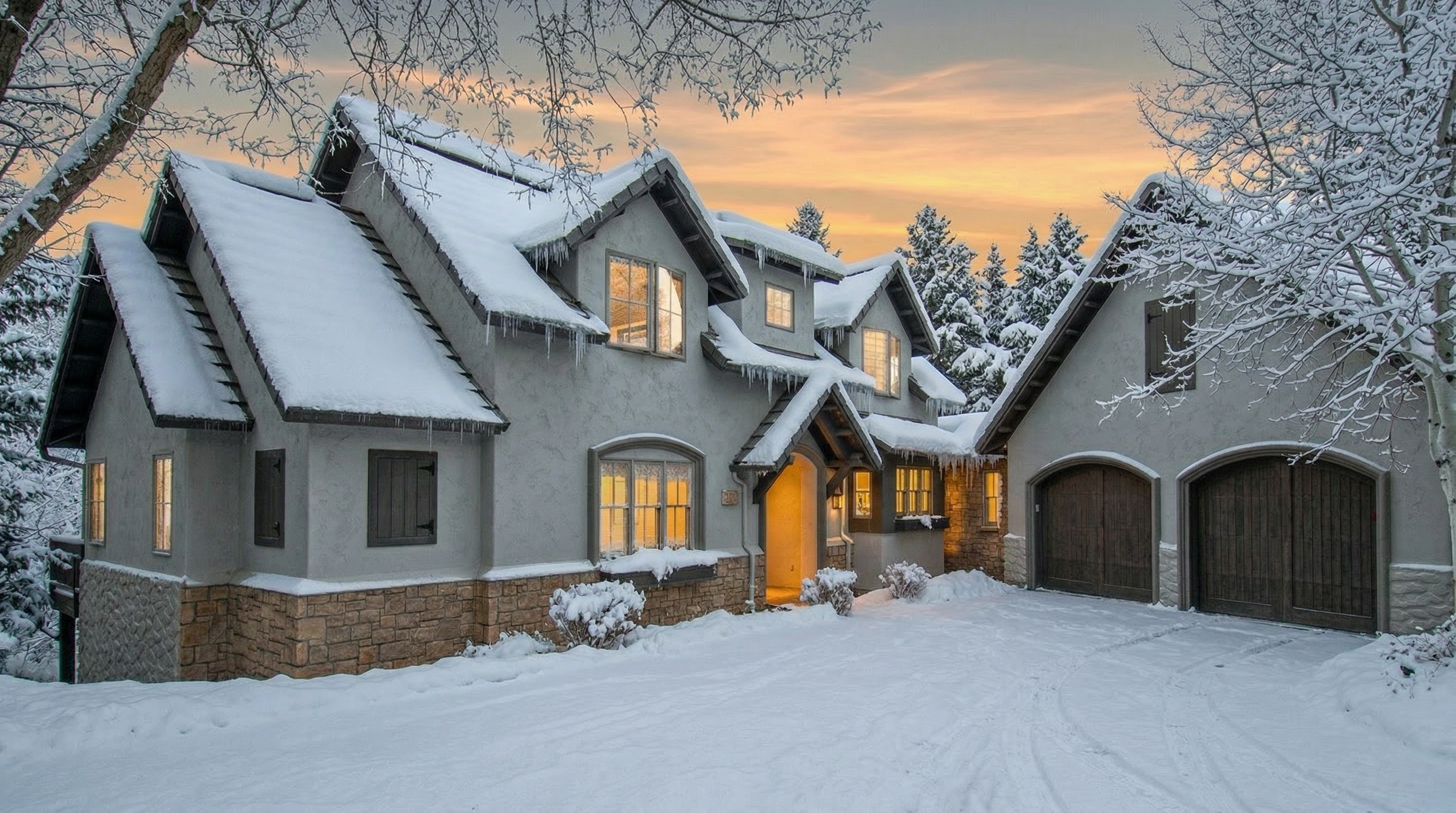 This charming, multi-gabled home features a classic exterior with a blend of neutral stucco and stone veneer masonry. Set against a picturesque winter backdrop with snow-covered trees and a dusting of snow, the residence creates an inviting atmosphere with warm, glowing interior lighting. The property includes a functional two-car side-entry garage and a welcoming entryway, projecting a cozy, upscale mountain-style aesthetic.