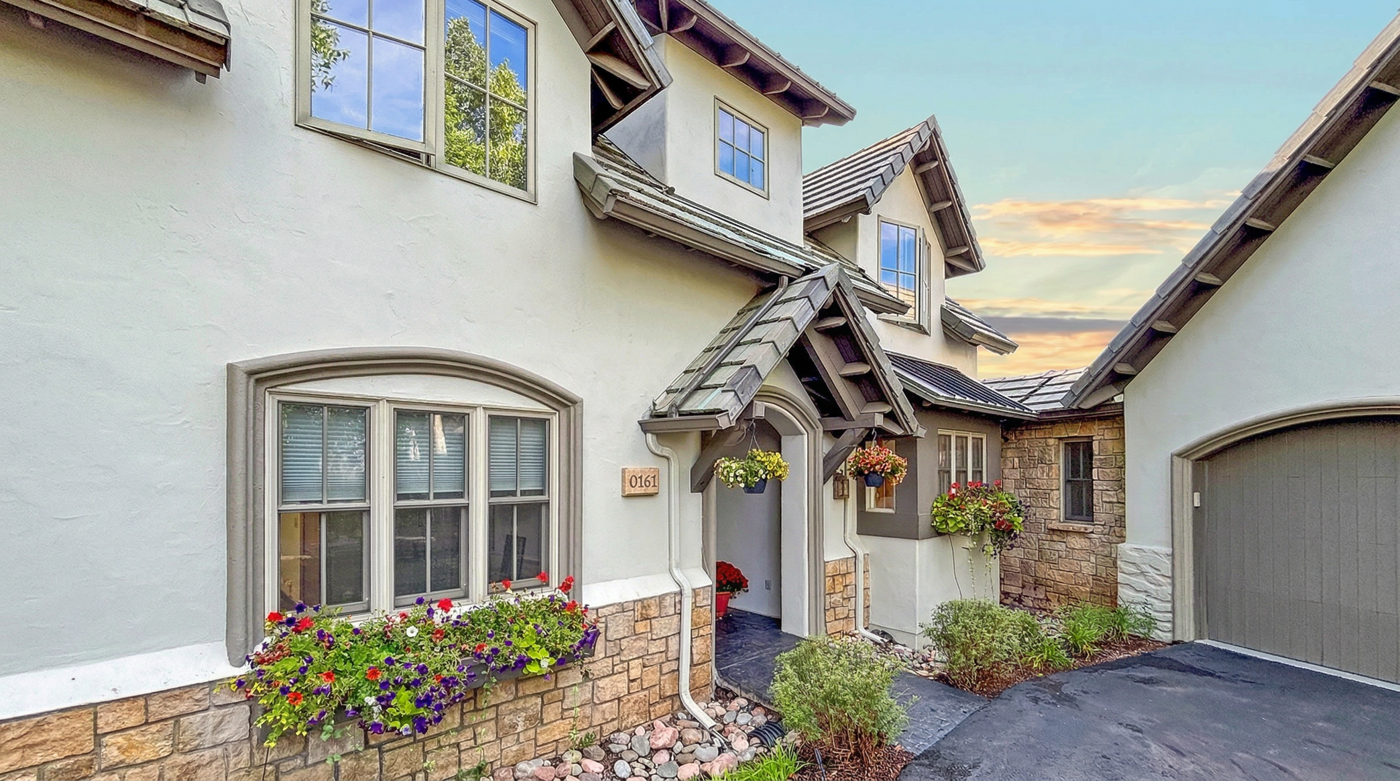 This charming home features a mix of stucco and cultured stone siding, highlighting a refined and welcoming curb appeal. Key details include a covered entryway adorned with hanging flower baskets, a large front window box filled with vibrant blooms, and an integrated garage. The overall architectural style suggests a tasteful, well-maintained residence with beautiful landscaping.