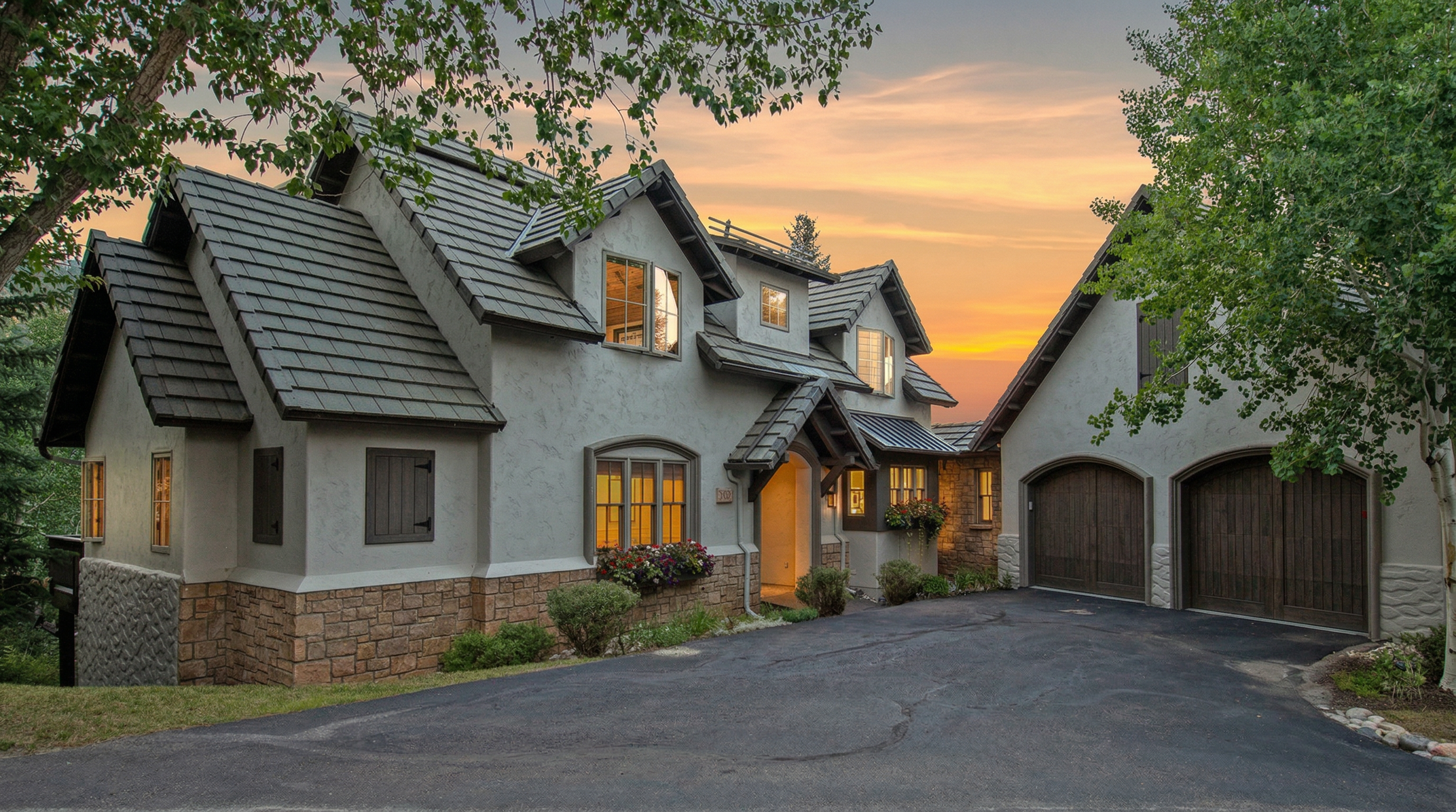 This stunning residence features an elegant, European-inspired exterior with a mix of stucco and natural stone cladding. Noteworthy elements include a multi-gabled roofline, decorative window shutters, and an attached two-car garage with arched wooden doors. Set against a dramatic sunset backdrop, the home's warm lighting and manicured flower boxes create a welcoming and premium impression.