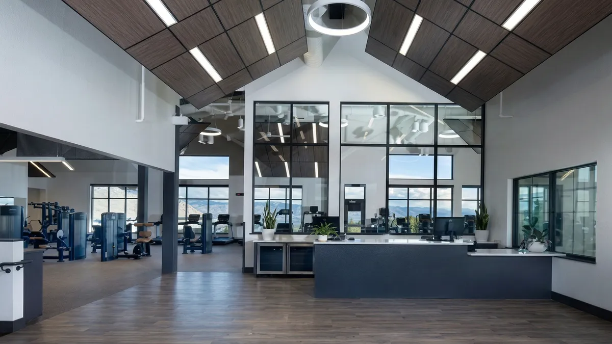 This professional-grade fitness facility features a sleek, modern design with a vaulted wood-paneled ceiling and recessed ambient lighting. The space includes a functional reception desk area, large windows that offer expansive mountain views, and a dedicated workout zone equipped with various exercise machines. The interior combines natural wood tones with sharp dark accents, creating an upscale and inviting atmosphere for residents.
