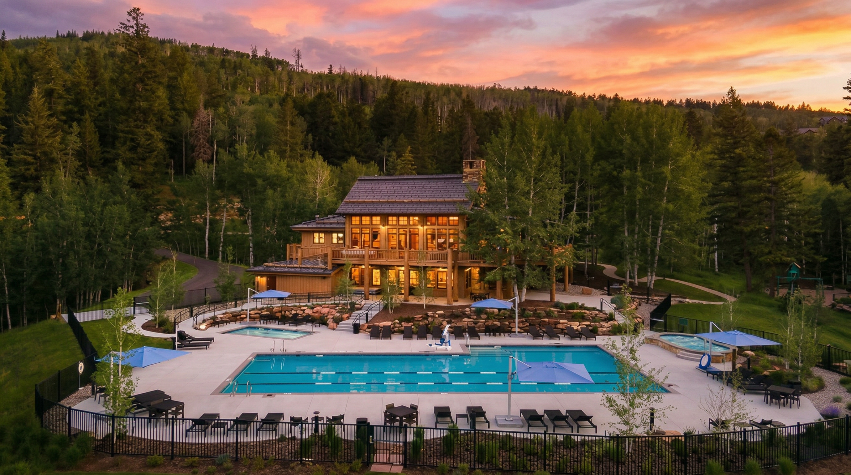 This scenic community amenity area features a large, lap-style swimming pool and a separate hot tub set against a backdrop of a luxurious mountain lodge. The property is nestled in a lush, forested environment at dusk, offering a relaxing retreat with ample patio seating and beautifully landscaped stone retaining walls. The rustic, timber-framed architecture of the lodge creates an inviting and upscale atmosphere for residents.