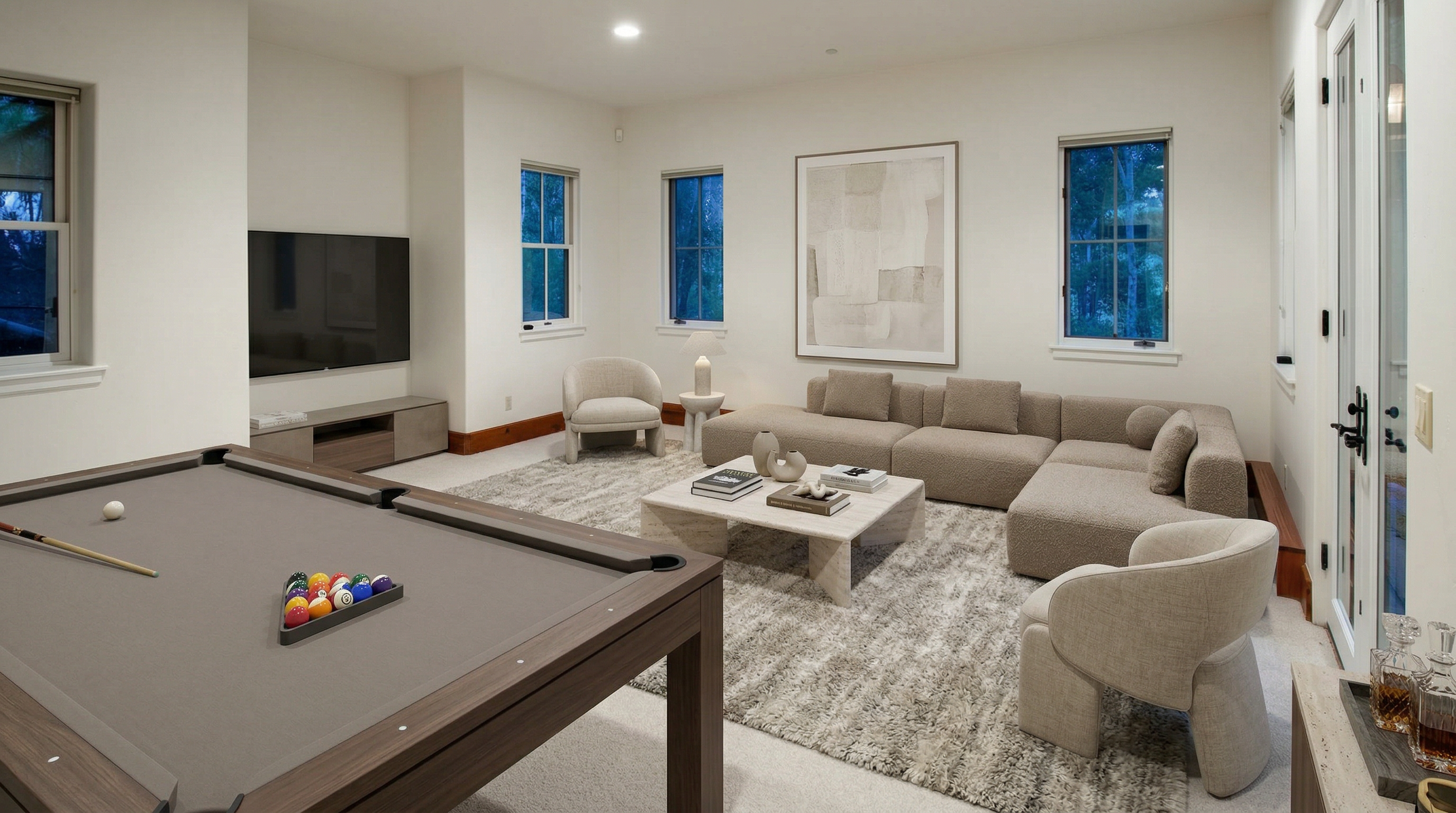 This spacious and modern living area features a clean, neutral color palette with plush textured upholstery and a sleek stone coffee table. The room serves as a multi-functional space, incorporating a home entertainment area with a large television and a sophisticated game room section complete with a pool table. Large windows and a glass door provide abundant natural light, creating an inviting, open-concept atmosphere.