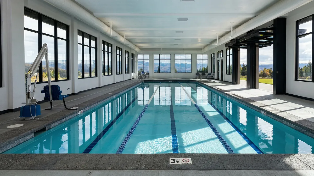 This facility features a bright, indoor lap pool with large windows that offer expansive mountain views. The space is modern and airy, incorporating a pool lift, lane markers, and a clean, minimalist design ideal for community exercise or relaxation. Large folding glass doors on the right open to the outdoors, providing a seamless transition between the indoor pool area and the exterior environment.