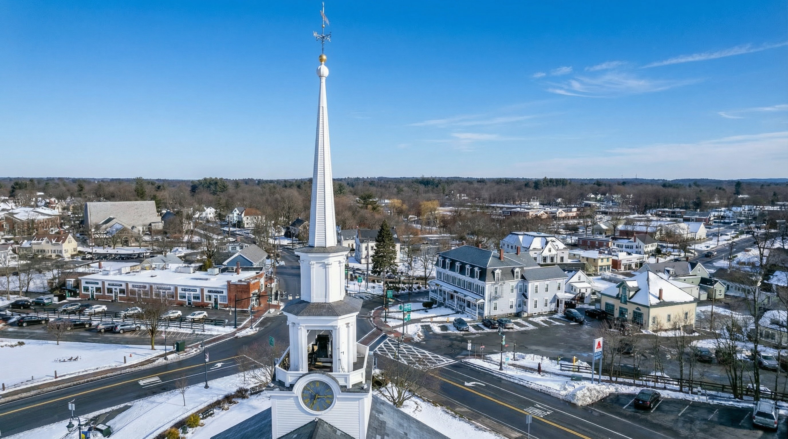 This aerial view showcases a quaint town in winter, dominated by a tall white church steeple with a visible clock. The surrounding buildings include residences and businesses, some with snow-covered roofs and parking areas. The image provides a comprehensive overview of the neighborhood's layout and architectural diversity.