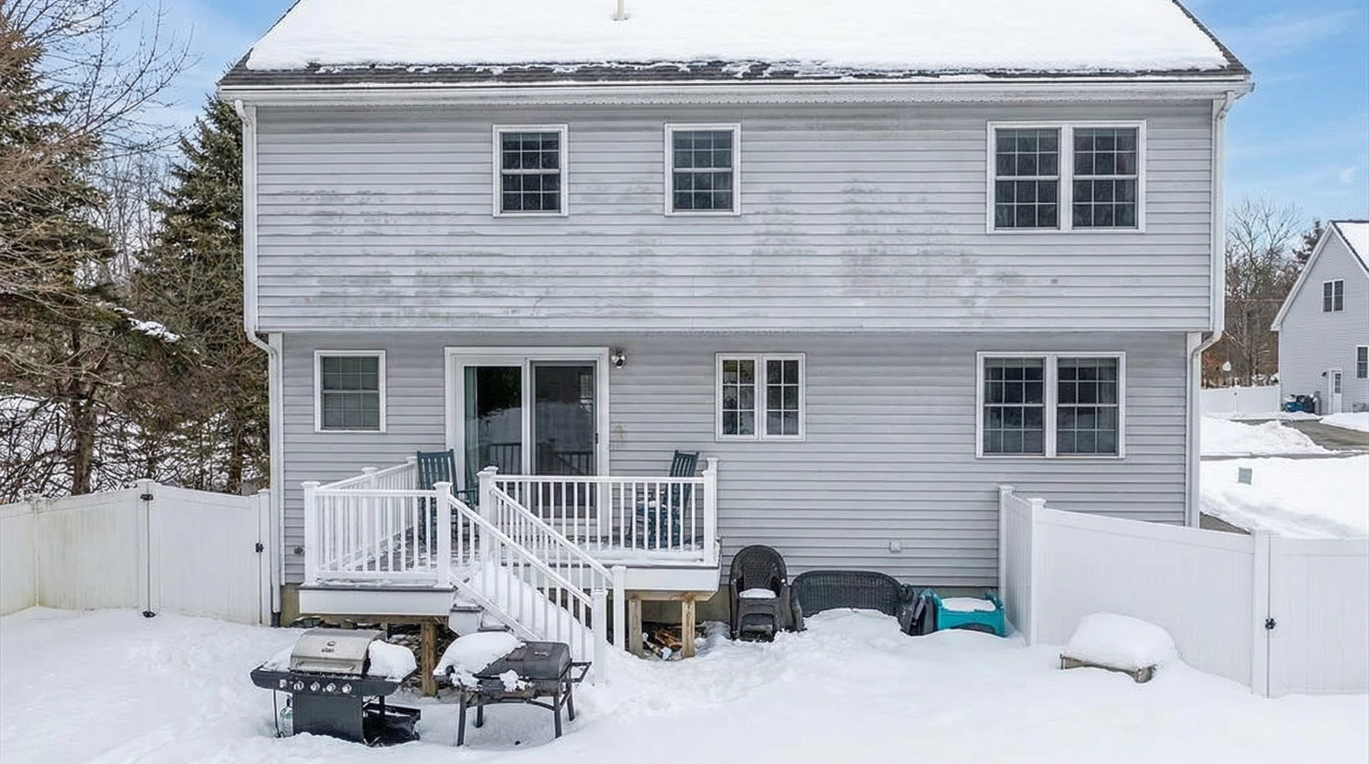 This image showcases the rear exterior of a two-story house with light gray siding. A deck with white railings and steps leads down to a snowy yard. The house features several windows and a sliding glass door on the first level.