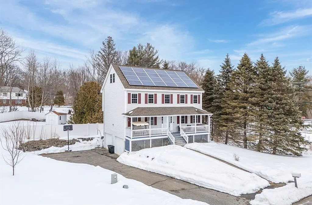 This is an exterior front view of a two-story home with white siding, red shutters, and solar panels installed on the roof. It features a covered front porch with white railings and steps leading up to the entrance. The surrounding landscape is partially covered with snow, with some trees and a driveway visible.