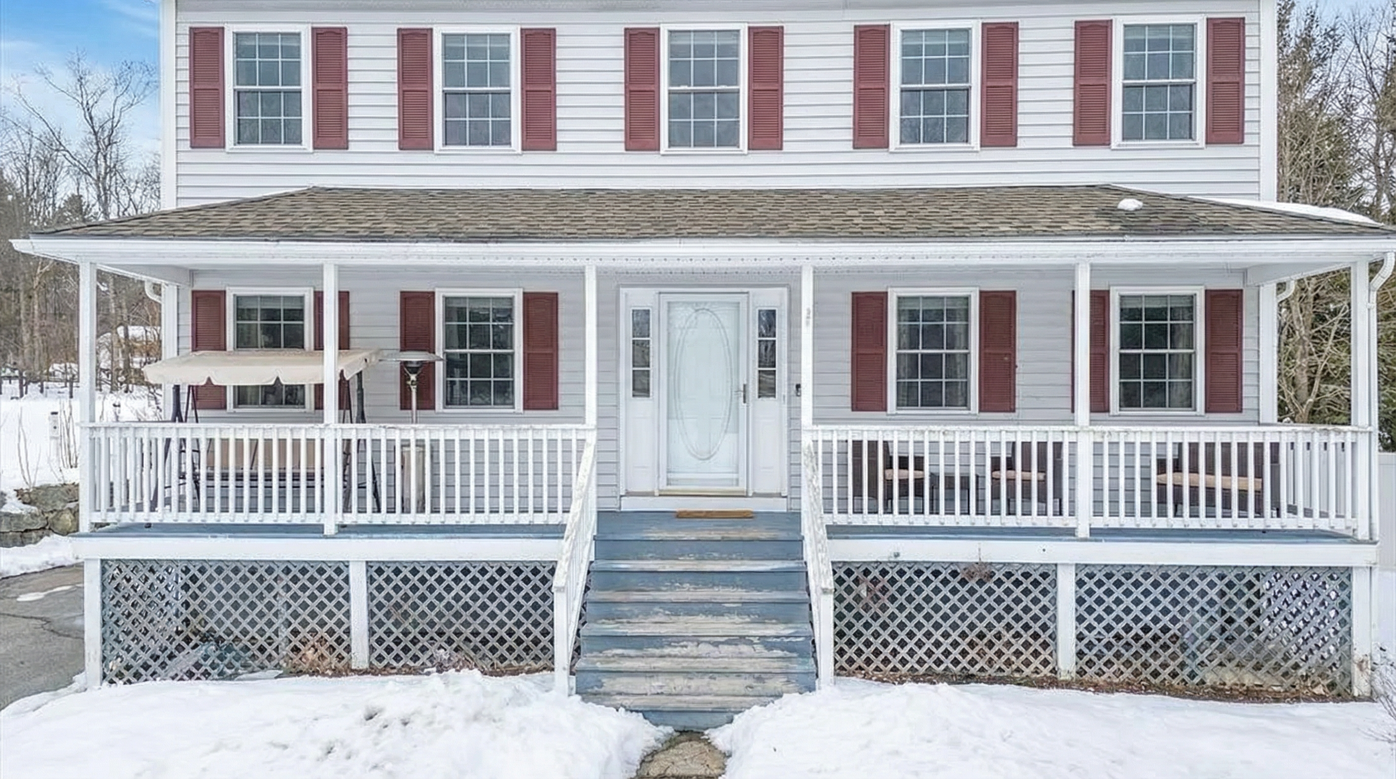 The image showcases the front exterior of a two-story home, emphasizing its welcoming facade. A covered front porch spans the width of the house, furnished with seating and supported by white columns, adding to the curb appeal. The house features white siding and contrasting red shutters, creating a classic and inviting appearance, despite the snowy foreground.