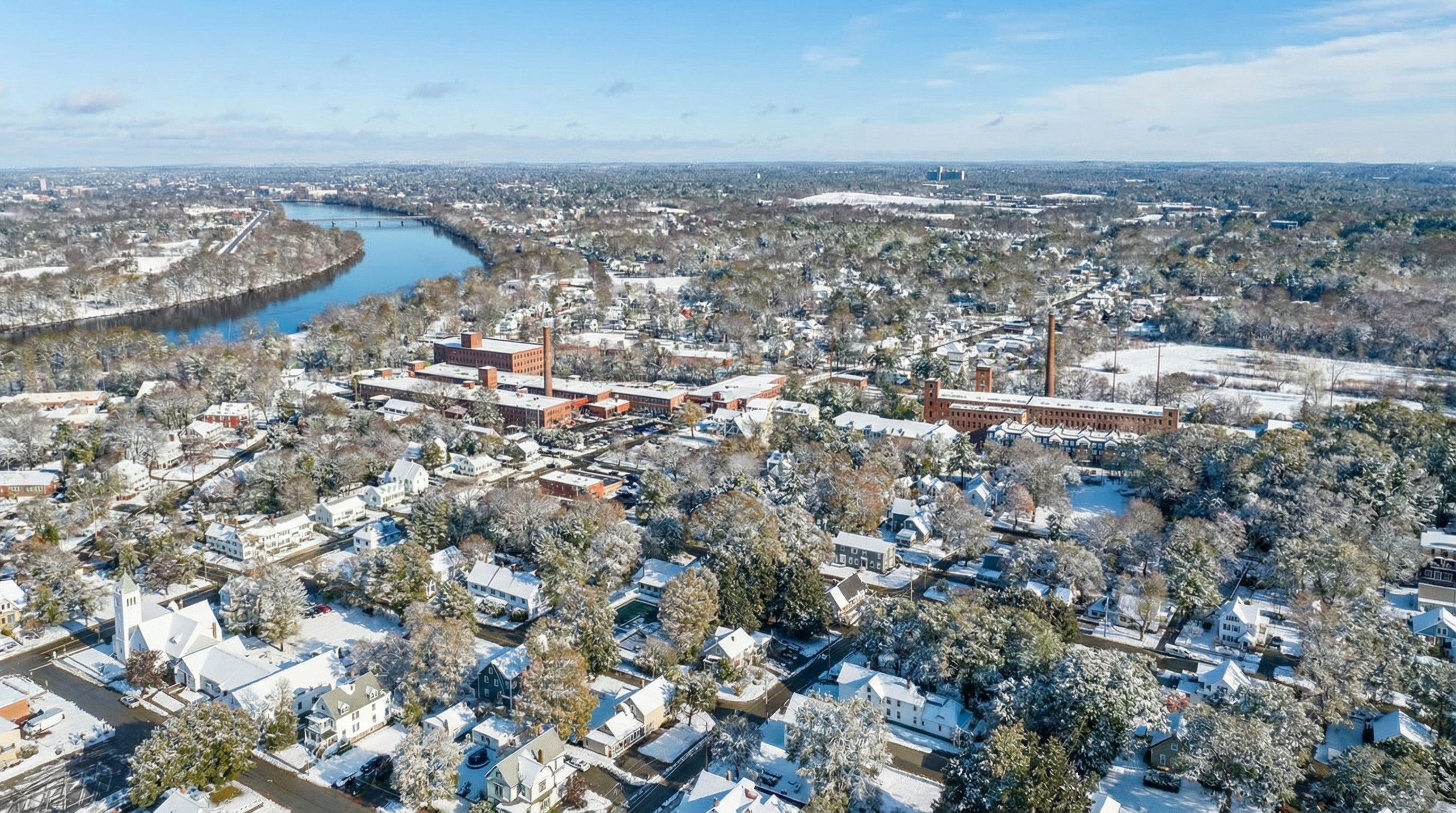 This aerial view showcases a snowy landscape featuring residential homes and an industrial area, likely a renovated mill or factory. The scene presents a blend of residential comfort and historical architecture, all blanketed by fresh snow. A river winds through the scene, adding a serene element to the wintery town.