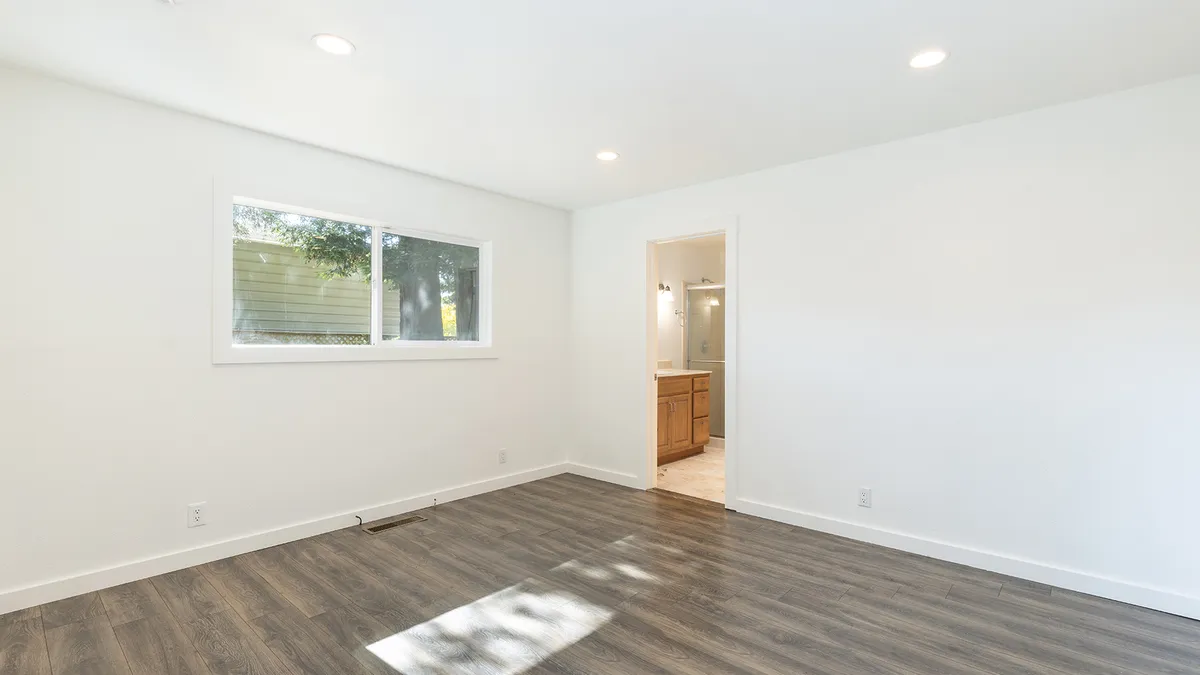 This spacious, modern bedroom features neutral white walls, recessed ceiling lighting, and low-maintenance wood-look vinyl plank flooring. A large horizontal window provides ample natural light, while a doorway leads directly into an en-suite bathroom, highlighting a seamless and functional layout suitable for a master suite.
