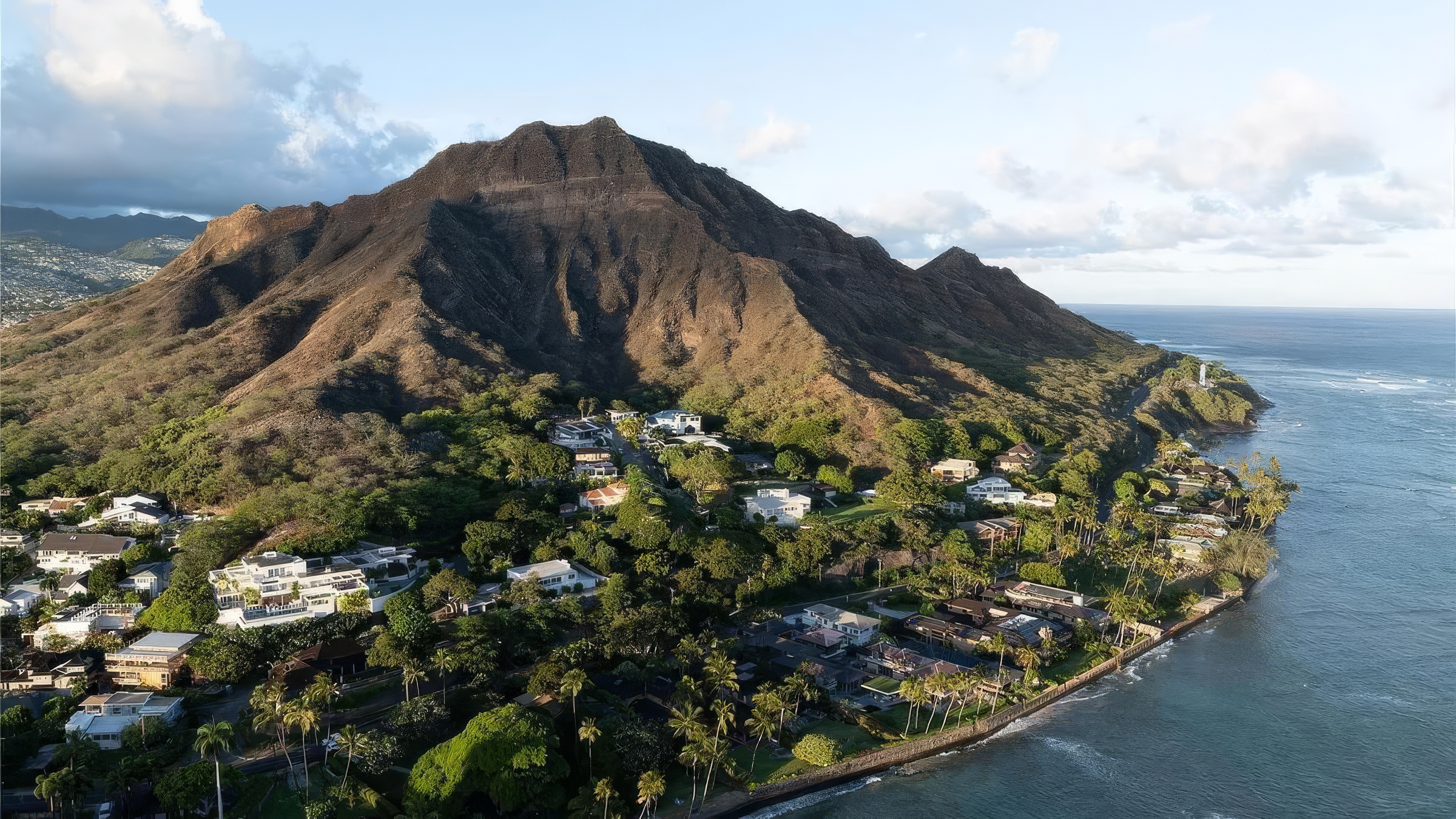 An aerial view showcases a neighborhood nestled along the coastline and at the base of a large mountain. The houses appear luxurious, featuring multiple levels, pools, and well-manicured landscaping with palm trees. The turquoise ocean provides a stunning backdrop, enhancing the property's appeal and highlighting the desirable coastal location.