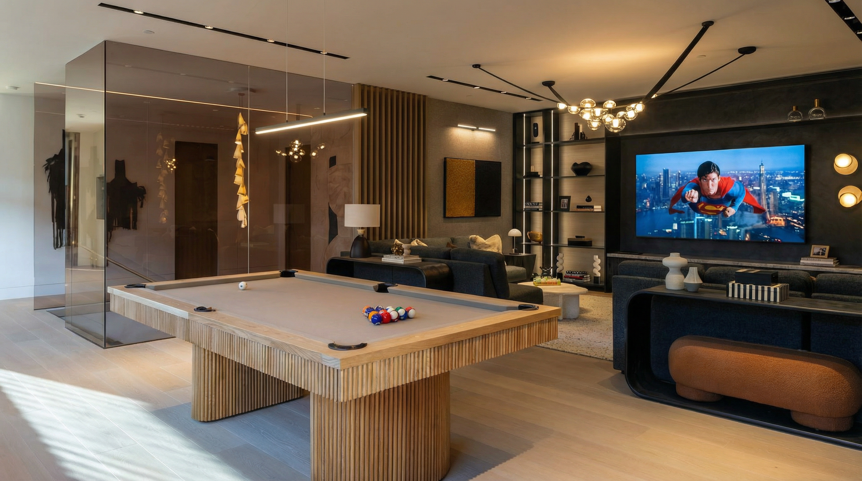 This interior shot showcases a modern living room, complete with a pool table and a large screen TV displaying Superman. The design incorporates a mix of textures, including a slatted wood feature behind the sofa and a unique, globe-pendant chandelier. Track lighting accentuates the space, and the overall impression is luxurious and inviting.