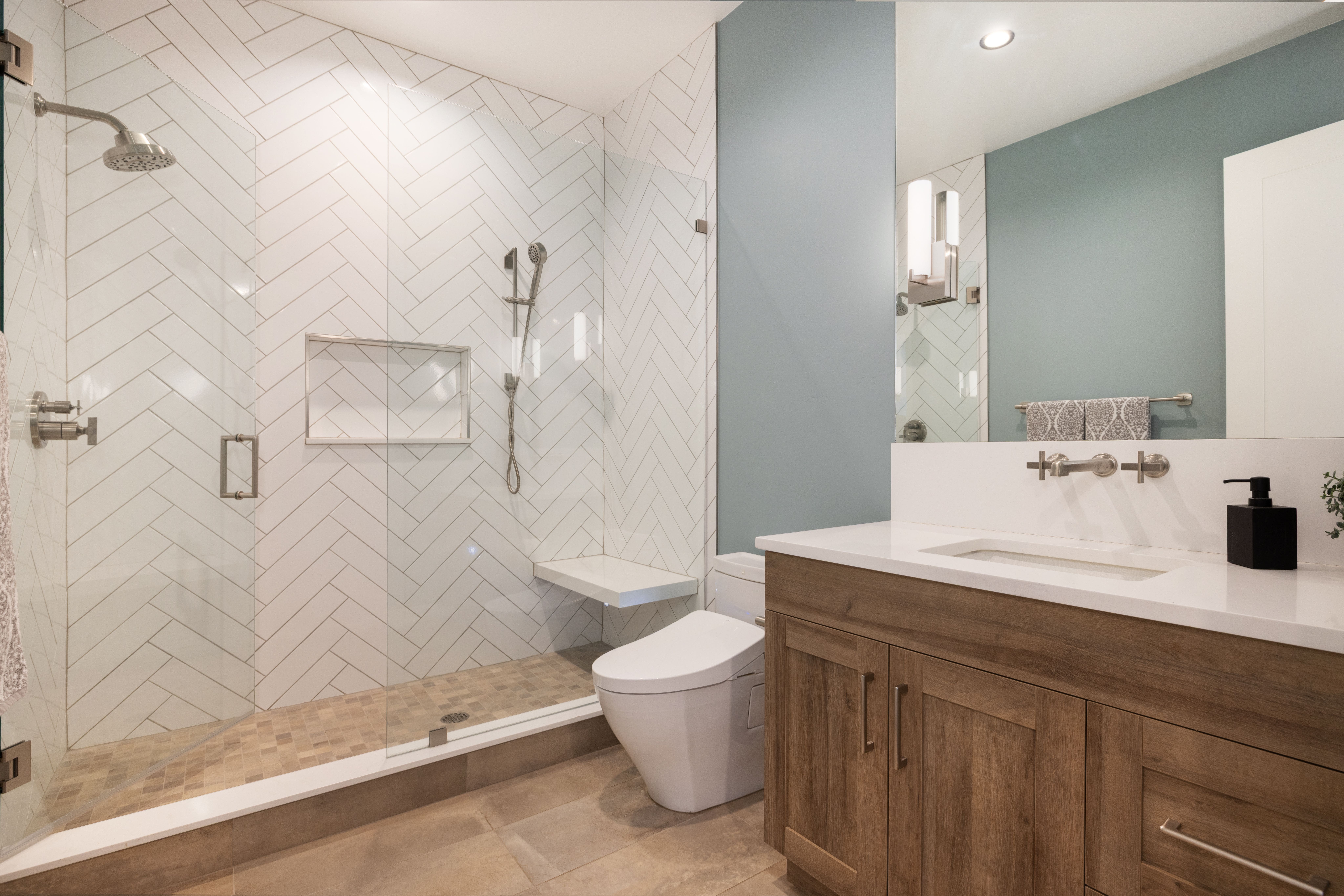 This modern bathroom features a spacious glass-enclosed shower with stylish herringbone-patterned wall tiles, a built-in bench, and a rainfall showerhead. The vanity, constructed with rich wood-toned cabinets and a white countertop, is complemented by a vibrant blue accent wall that adds a refreshing splash of color. The space is well-designed, blending functional storage and elegant fixtures for a sophisticated, contemporary feel.