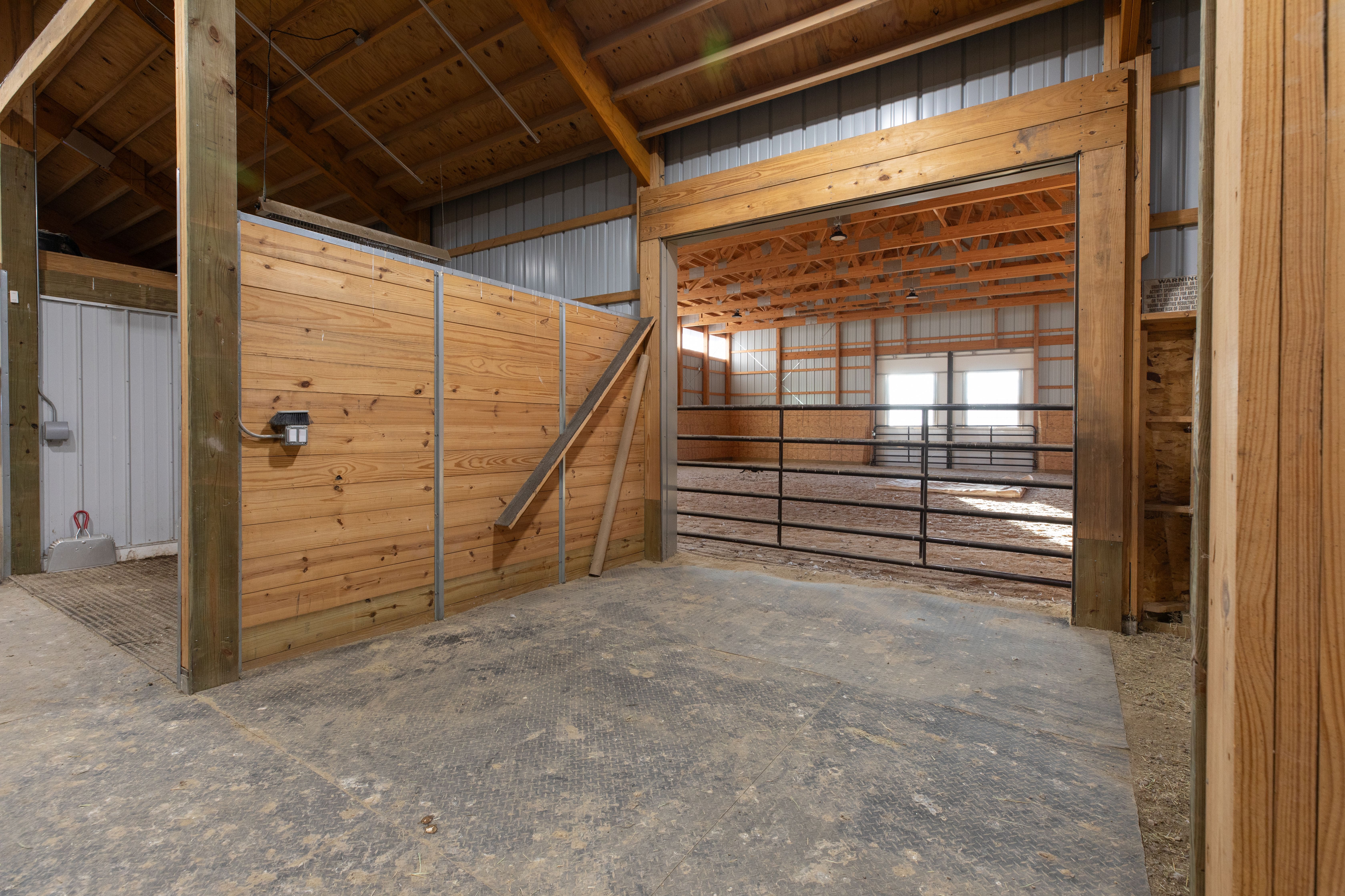 This image shows the interior of a horse stable or indoor riding arena, featuring timber framing and metal siding. The foreground highlights a stable stall with wooden partitioning, while the background reveals a large, open equestrian ring with metal fencing. The space has a rugged, functional aesthetic characteristic of agricultural facilities.