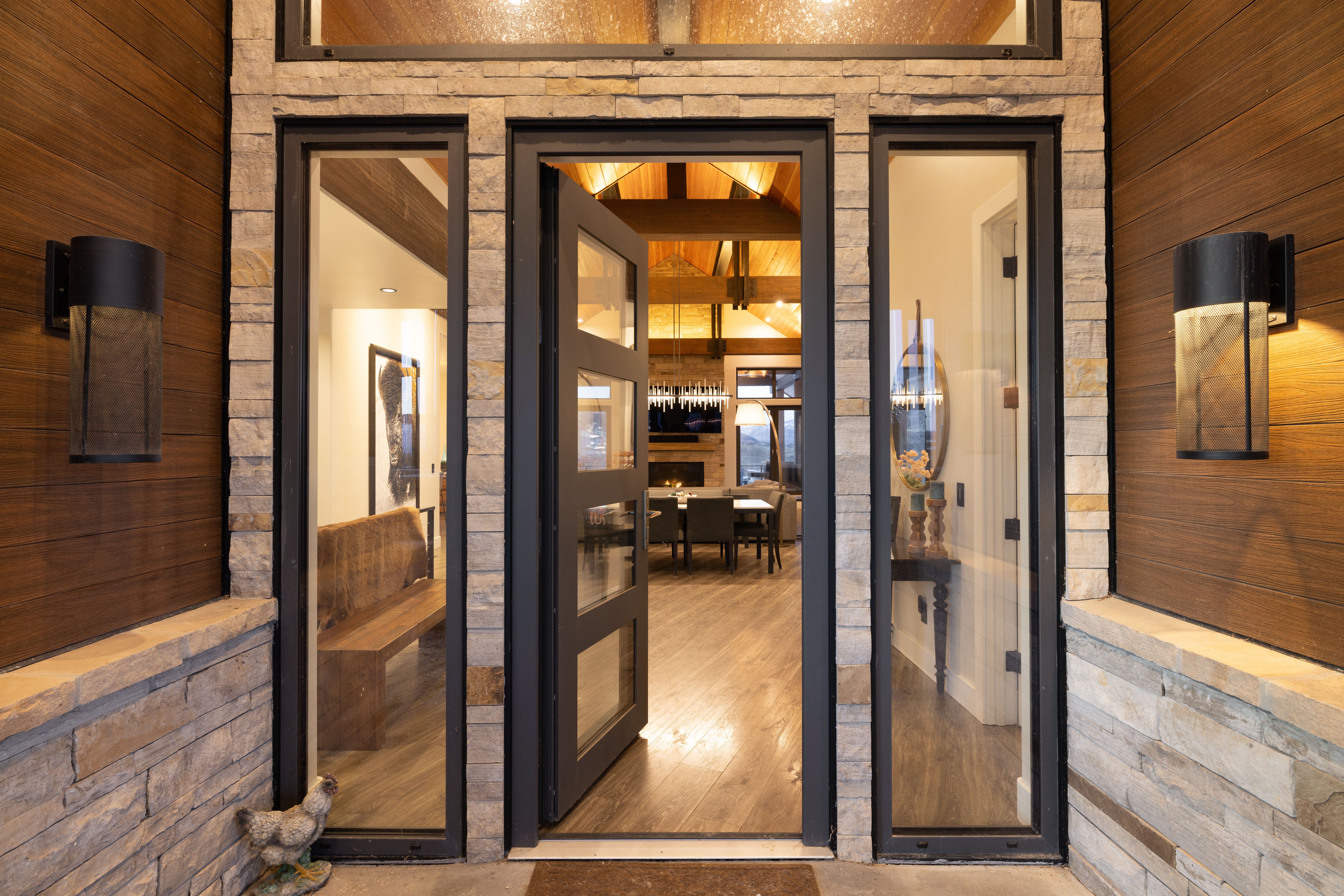 This striking entrance features a modern door with glass panels framed by natural stone masonry and warm wood siding. Matching cylindrical sconces with a unique mesh design provide elegant outdoor lighting, while the clear view through the entryway reveals a sophisticated and inviting home interior. The architectural blend of stone and wood creates a high-end, contemporary mountain aesthetic.