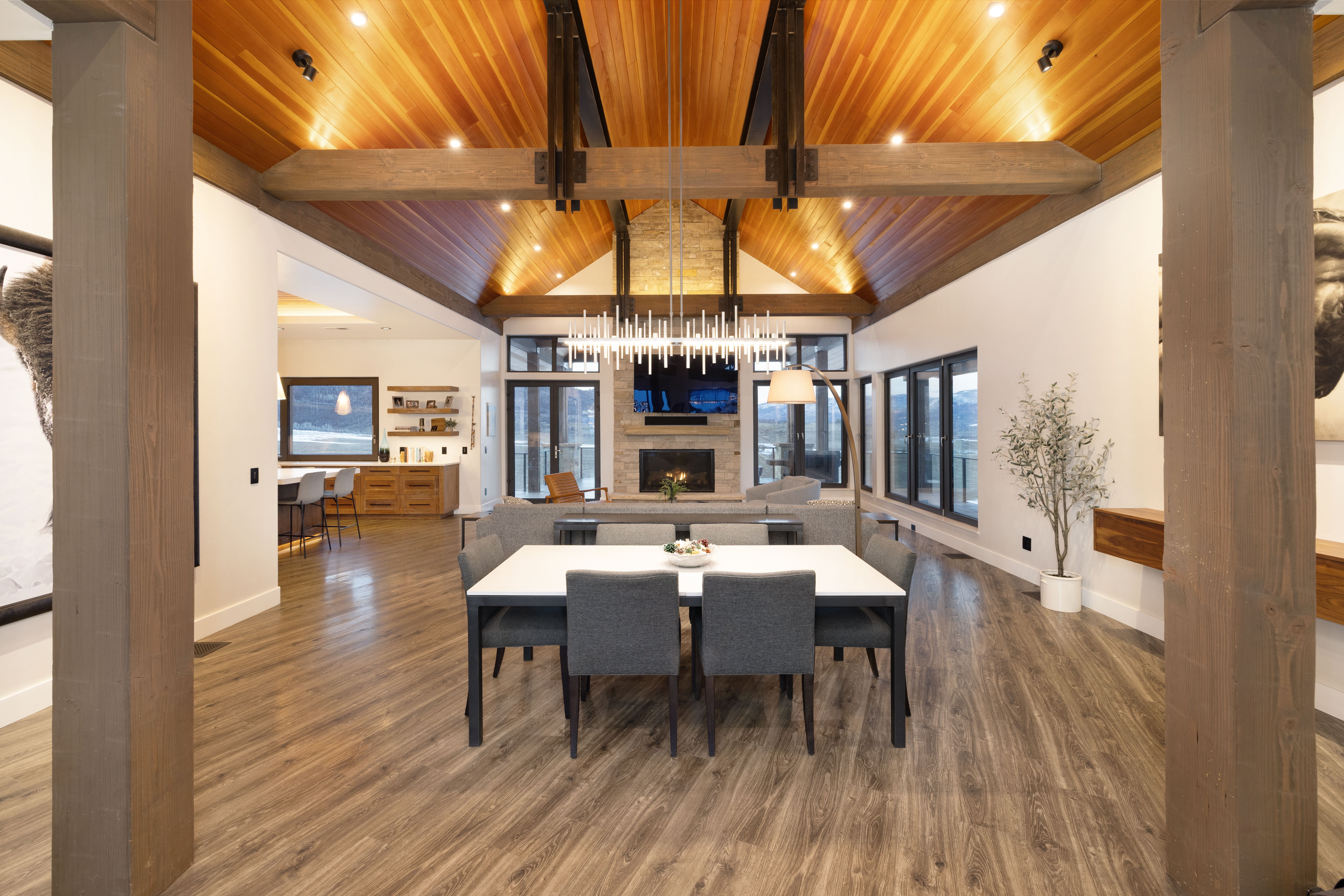This spacious open-concept home features a sophisticated dining area that flows seamlessly into a comfortable living room with a large stone fireplace. The vaulted ceiling with exposed wood beams and warm wood paneling creates a grand, rustic-modern atmosphere, complemented by high-end finishes and clean lines. An adjacent kitchen with bar seating and large windows framing scenic views further enhances the appeal of this expansive, well-lit living space.