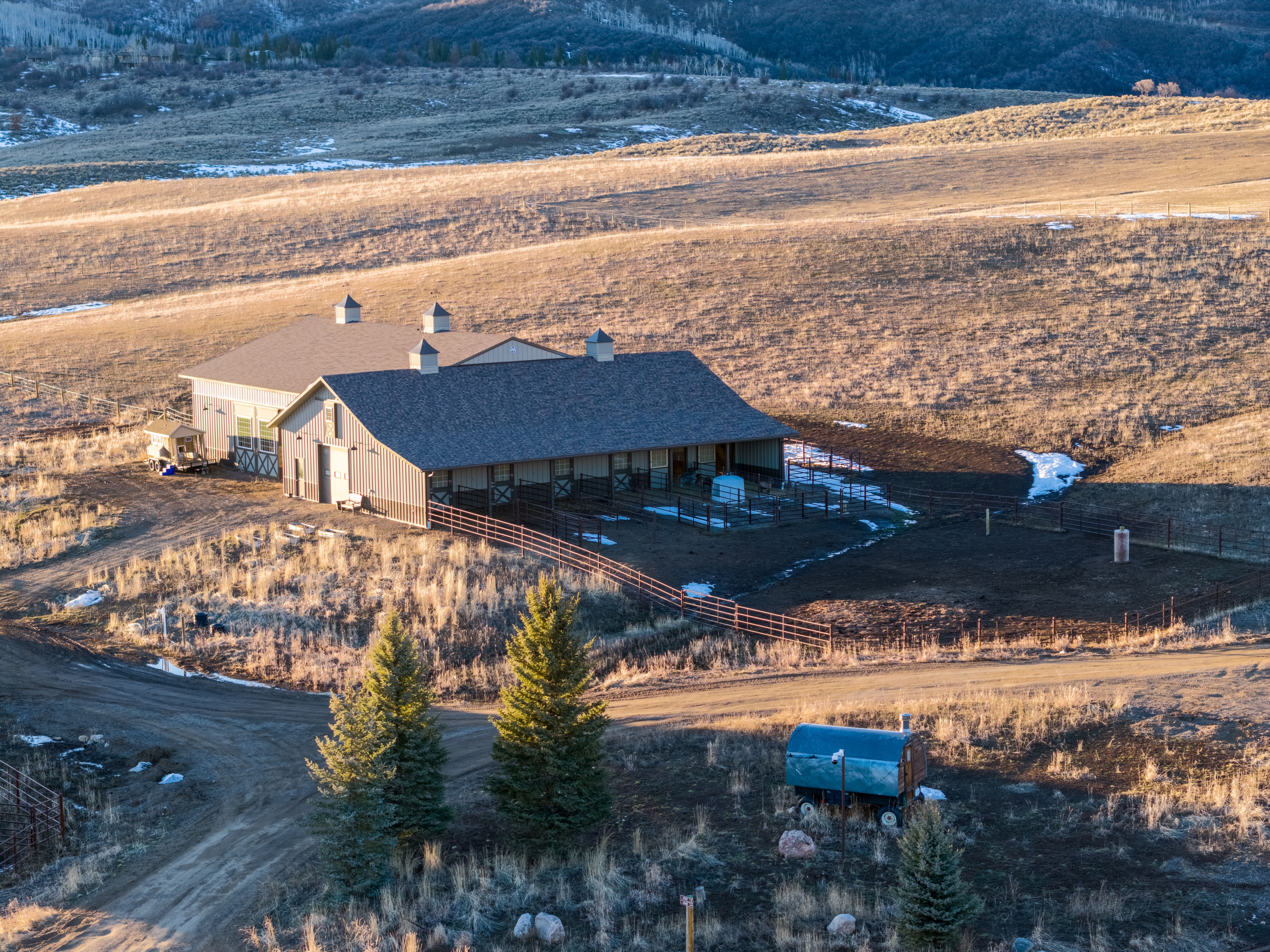 This high-angle, aerial perspective features a substantial, rustic barn structure situated in a vast, rolling mountainous landscape. The property includes fenced outdoor paddocks, a durable metal-sided exterior, a pitched shingle roof with decorative cupolas, and a charming, colorful wagon parked nearby. Its remote, scenic location and functional equestrian-style design offer a tranquil retreat ideal for hobbyists or livestock enthusiasts.