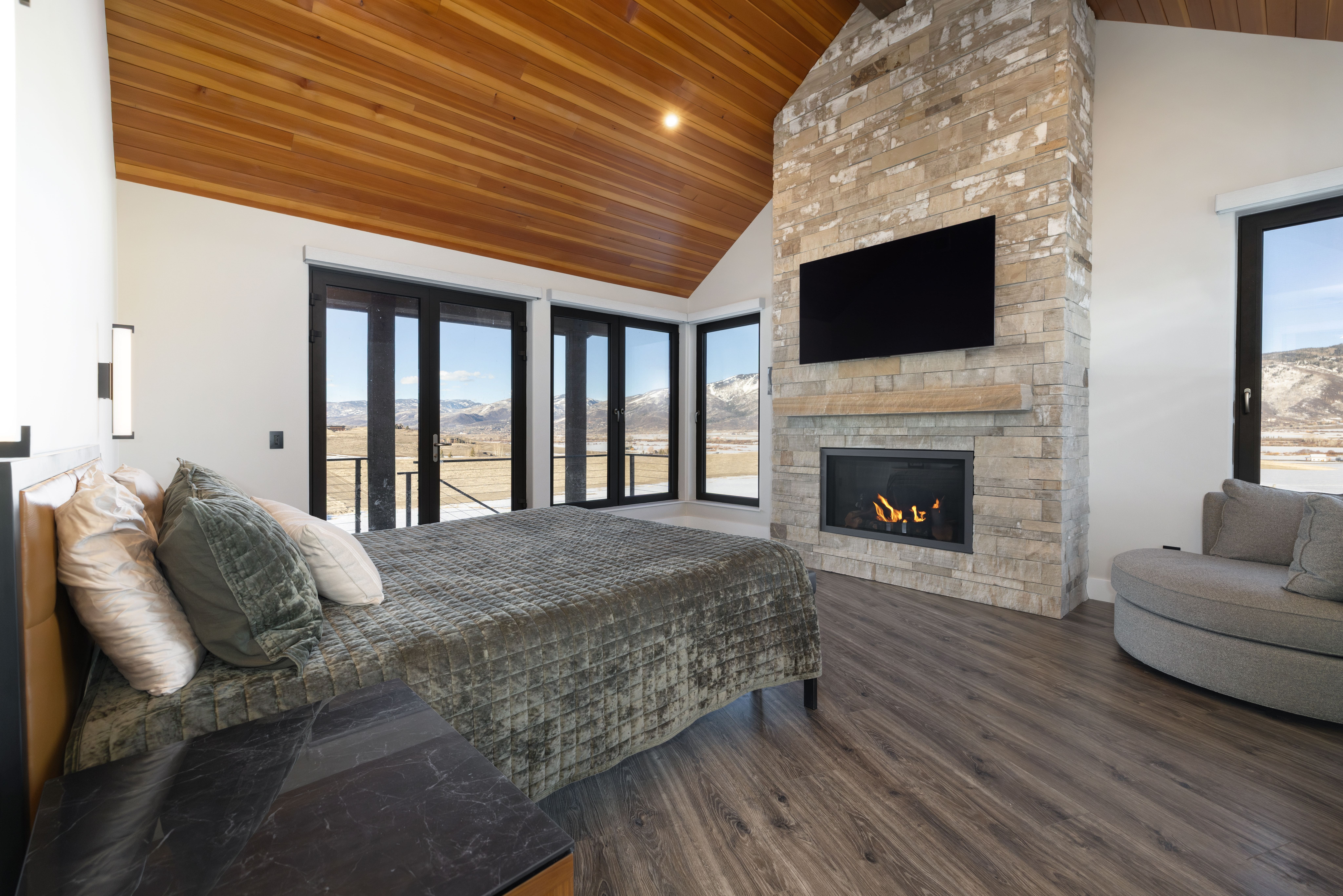 This elegant primary bedroom features a vaulted wood-paneled ceiling and a striking floor-to-ceiling stone fireplace as a central focal point. Large black-framed windows and doors offer expansive, scenic mountain views, while the room is anchored by a comfortable bed with plush green bedding and a contemporary grey accent chair. The space combines rustic stone elements with modern wood finishes, creating a sophisticated and luxurious bedroom interior.