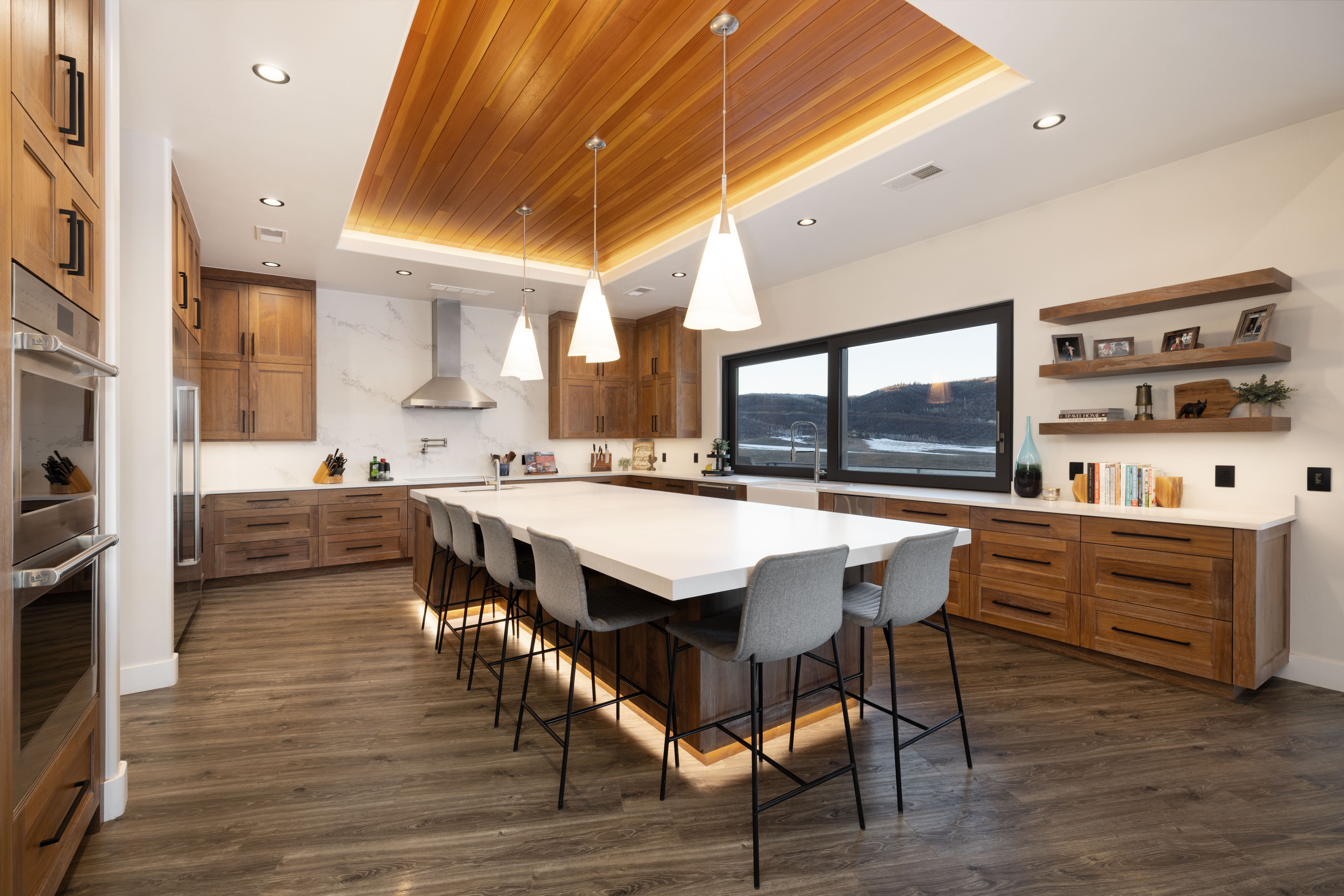 This stunning contemporary kitchen features high-quality wood cabinetry, a large white quartz island with barstool seating, and a unique wood-slat ceiling with integrated lighting. The space is equipped with modern stainless steel appliances, a sleek range hood, and strategically placed floating shelves against a warm, neutral wall. Large windows provide beautiful panoramic views, enhancing the bright and luxurious atmosphere of this well-designed home culinary space.