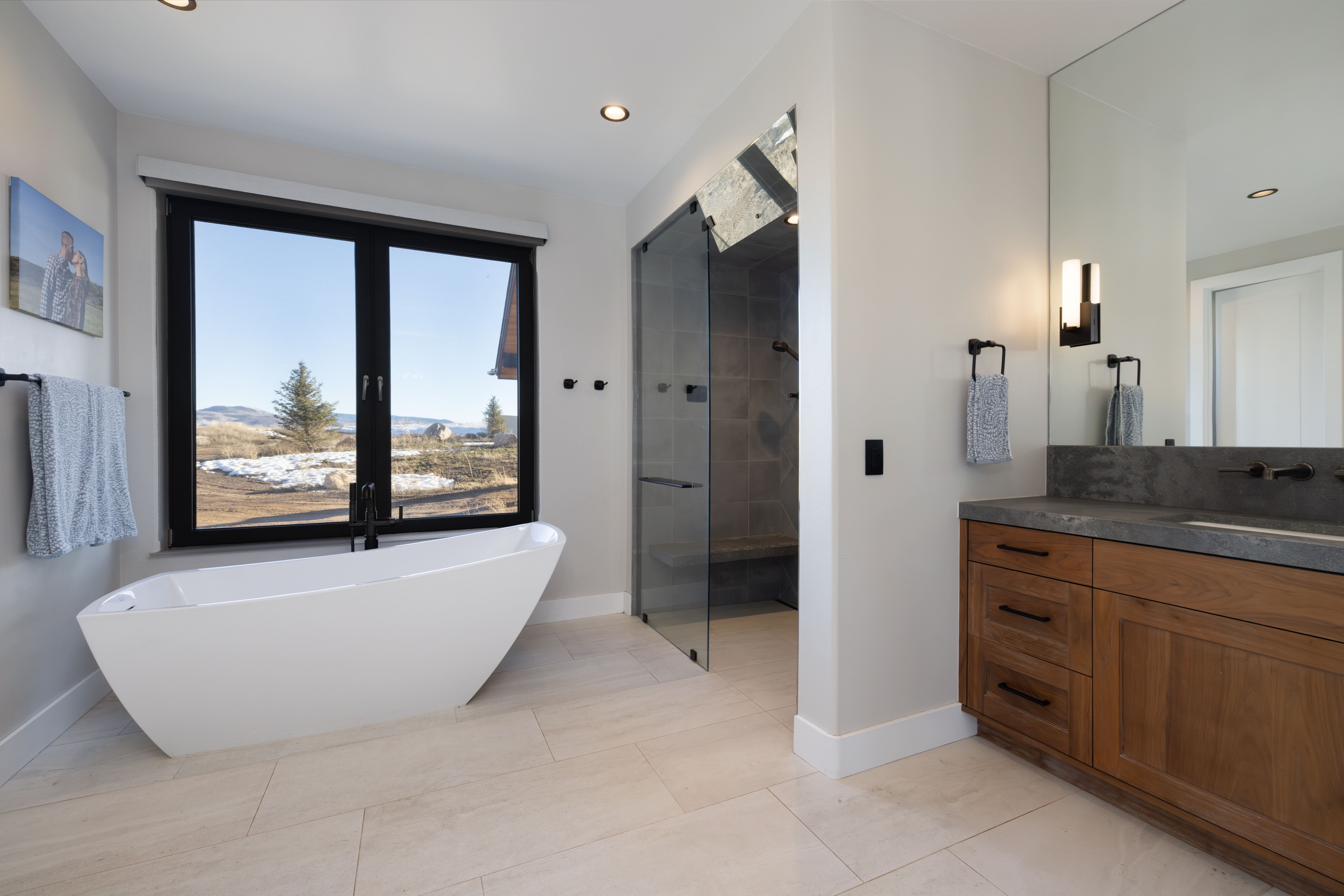 This modern primary bathroom features a sleek freestanding soaking tub placed by a large window with scenic outdoor views. It includes a walk-in glass-enclosed shower with gray tiling and a custom vanity with warm wood cabinetry and a dark stone countertop. The space is bright and airy, blending natural elements with contemporary fixtures for a spa-like feel.