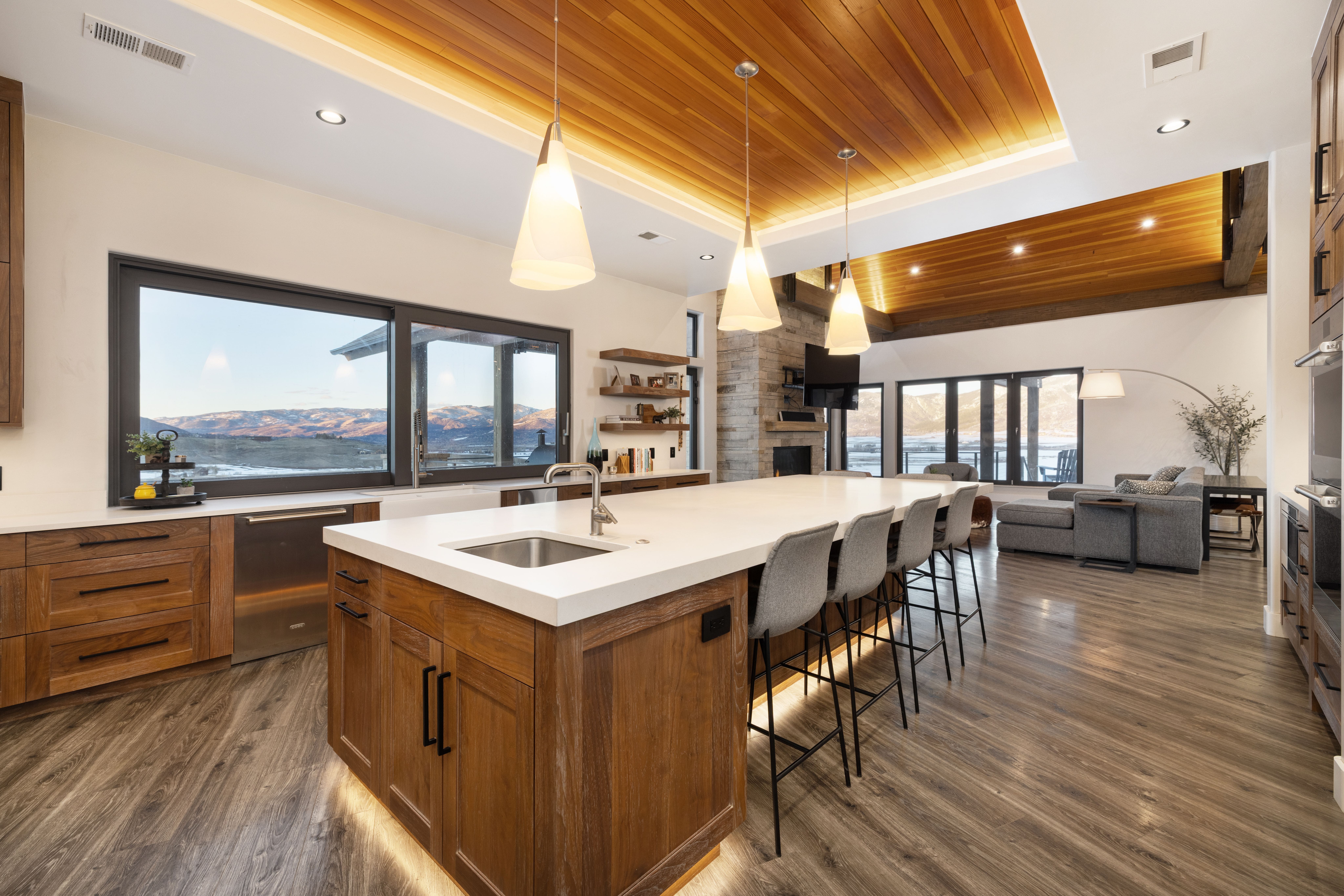 This stunning, open-concept kitchen features warm wood cabinetry, a large central island with quartz countertops, and modern gray seating. The space boasts a unique coffered wood ceiling, under-cabinet and under-island lighting, and expansive windows that frame breathtaking mountain views. It seamlessly integrates style and functionality, leading visually into a comfortable and bright living area.