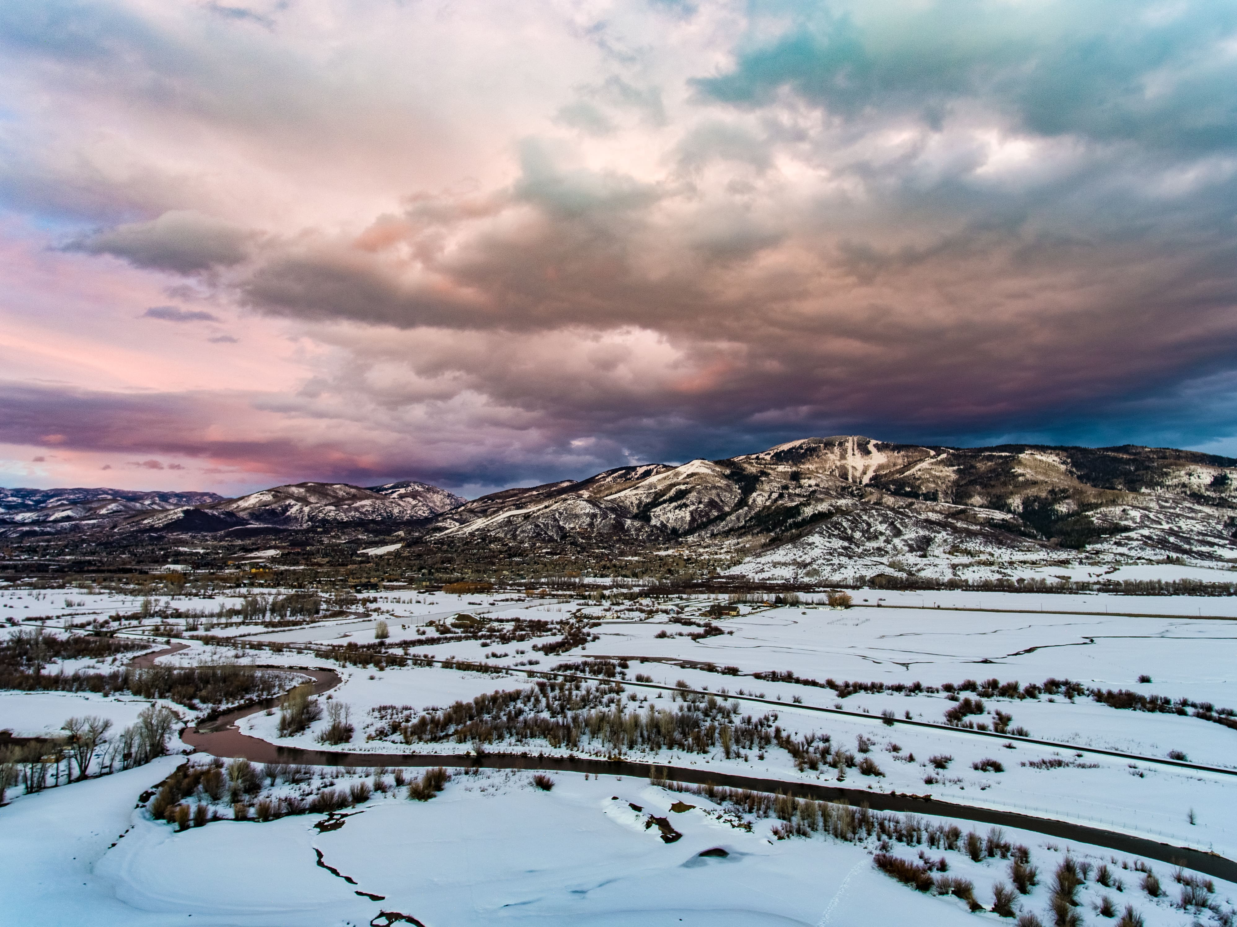 This panoramic aerial view captures a scenic mountainous landscape dusted with snow, featuring a prominent ski resort on the mountainside. A meandering river flows through the foreground of the snowy valley, highlighted by the soft, dramatic light of a cloudy sunset. The vast, open surroundings offer a picturesque and tranquil setting ideal for a mountain retreat.