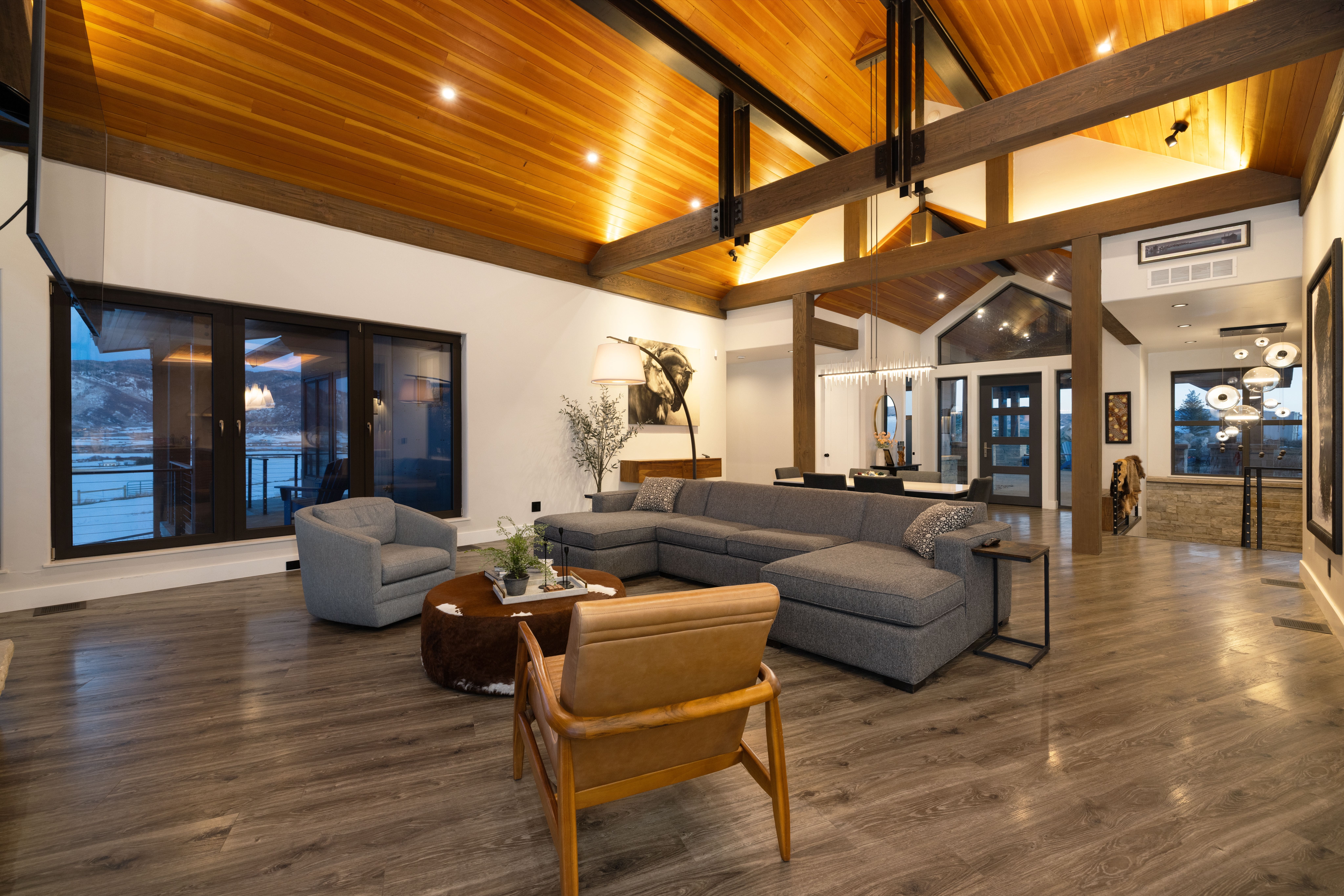 This spacious, open-concept living area features a warm wood-plank vaulted ceiling with striking iron structural beams and modern recessed lighting. The room is anchored by a large, comfortable gray sectional sofa, a brown leather armchair, and a circular ottoman, set against expansive windows that provide stunning mountain views. High-end finishes, neutral tones, and tasteful contemporary art create an inviting and sophisticated atmosphere, perfectly blending rustic charm with modern luxury.