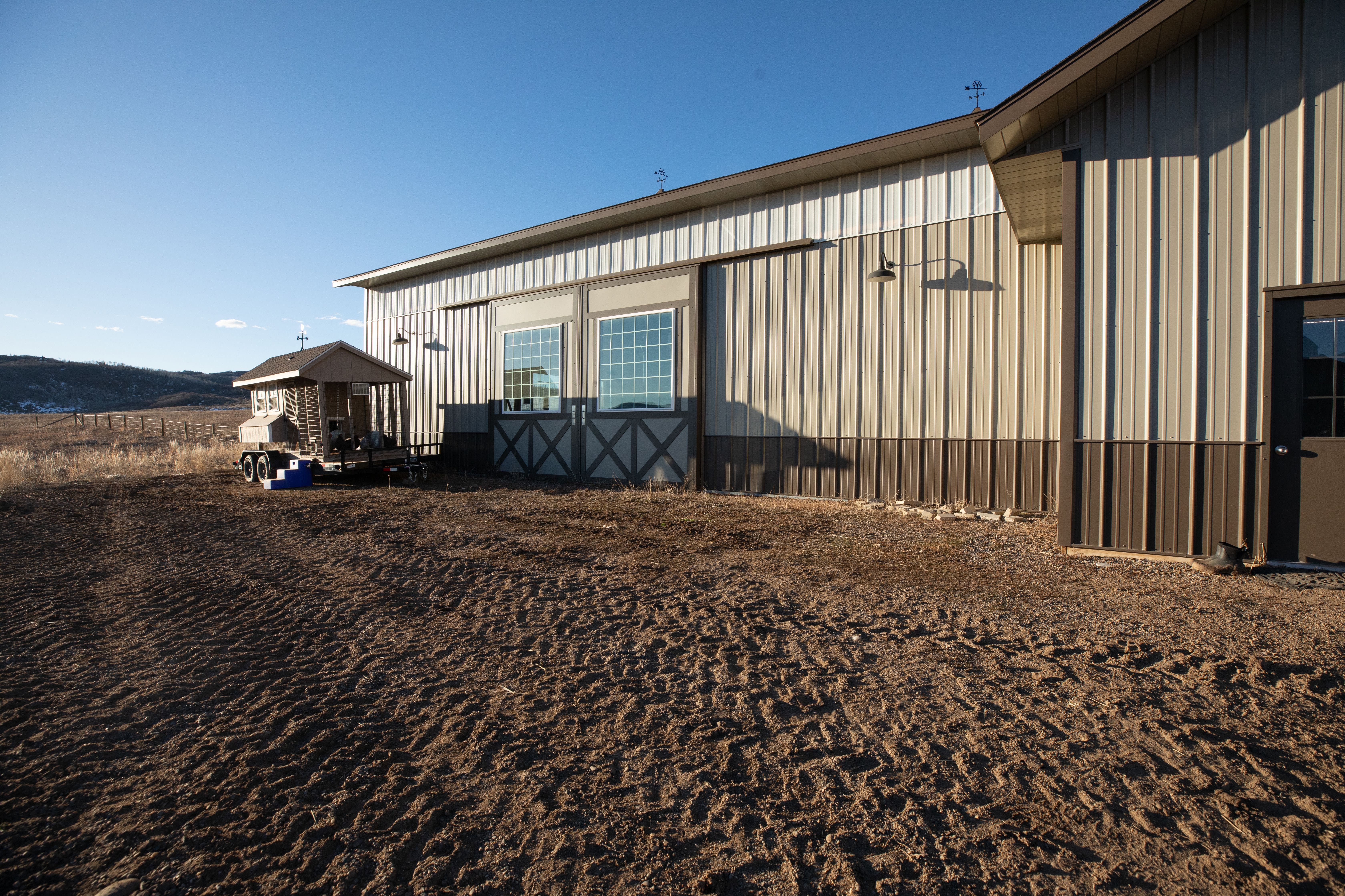 This exterior side view showcases a large metal-sided building with sliding barn-style doors featuring decorative cross-bracing and large windows. To the side, a portable chicken coop is parked on dry, uneven terrain in a rural, open landscape setting. The structure has a clean, utilitarian aesthetic, highlighted by industrial lighting fixtures and a neutral brown and cream color palette.