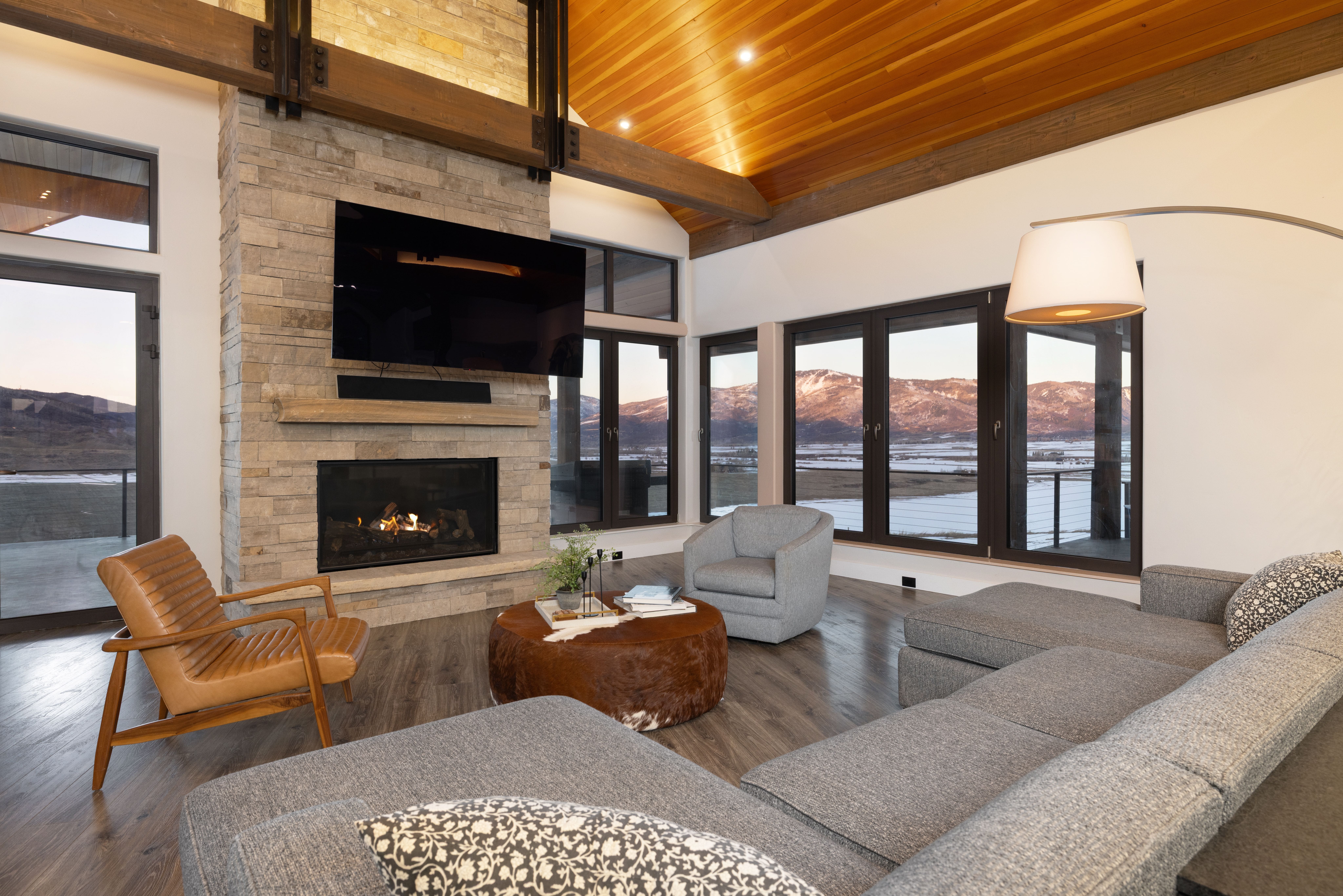 This spacious, modern-rustic living room features a striking floor-to-ceiling stone fireplace as a central focal point, complemented by high vaulted ceilings with natural wood cladding. The space is anchored by a large, comfortable gray sectional and a unique, circular cowhide coffee table, all set against a backdrop of expansive windows offering breathtaking mountain and valley views. The blend of warm wood tones, polished stone, and stylish furniture creates a sophisticated yet cozy atmosphere perfect for high-end mountain living.