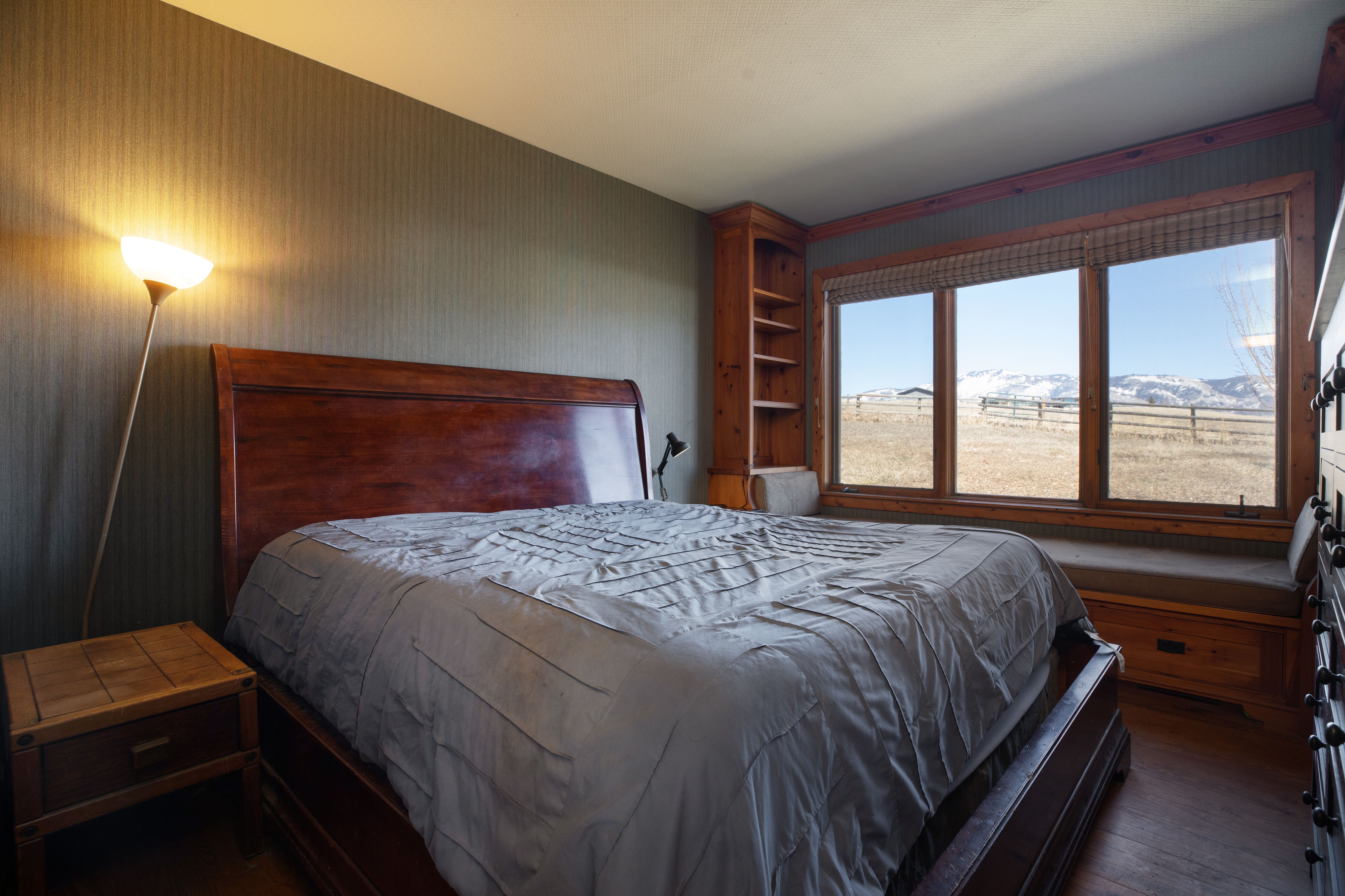 This primary bedroom features a classic wooden sleigh bed with a gray ruched duvet, complemented by a rustic wooden bedside table and a tall floor lamp. The room includes built-in wooden shelving and a window seat that overlooks a scenic, snow-dusted landscape, creating a warm, mountain-lodge aesthetic. The earthy color palette and textured wallpaper contribute to a cozy and inviting atmosphere.