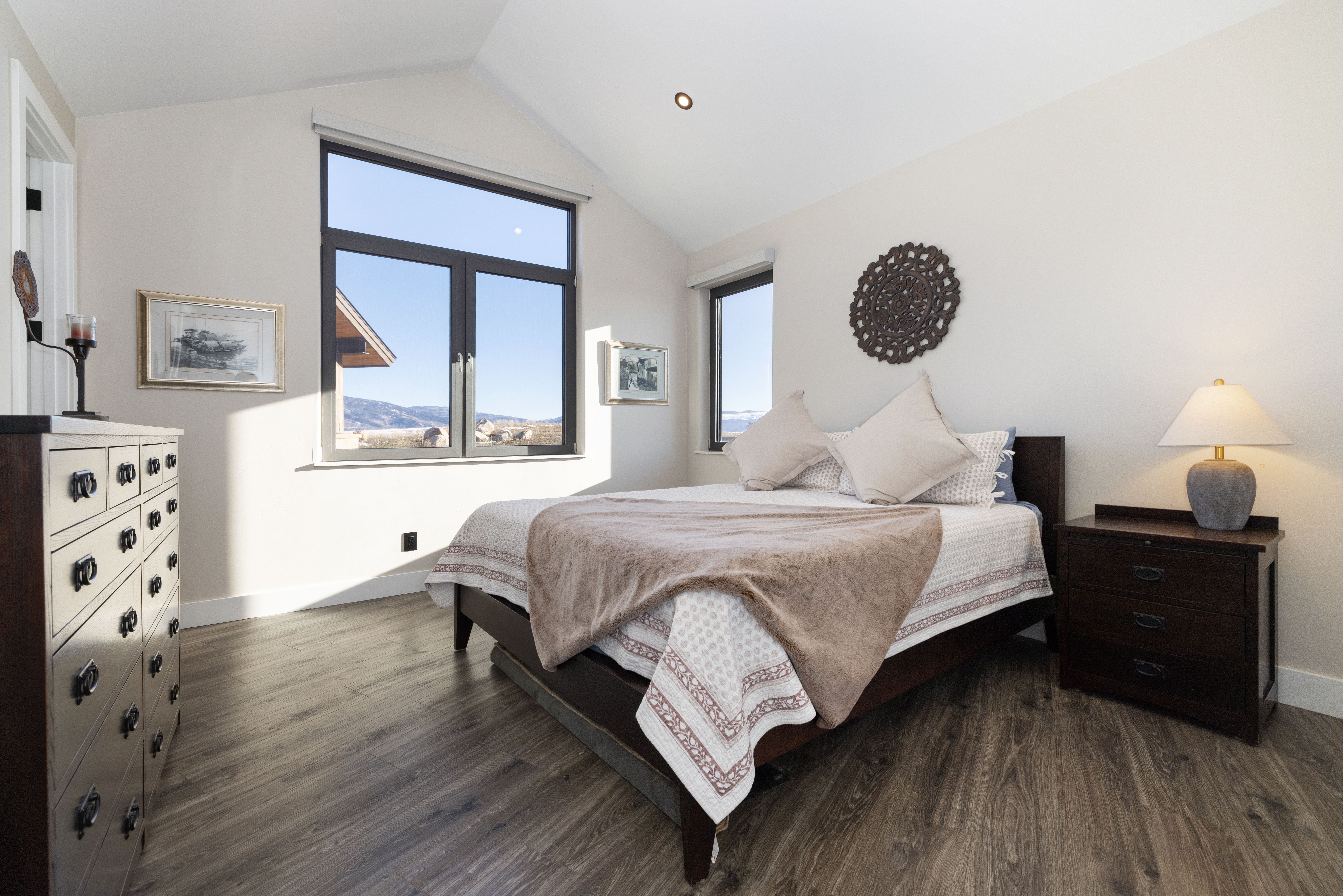 This serene primary bedroom features a modern aesthetic with vaulted ceilings, dark wood furniture, and high-quality wood-look flooring. Natural light floods the space through large, expansive windows that offer picturesque views of the surrounding mountainous landscape, while neutral bedding and an ornate wall decor piece add texture and comfort to the room.