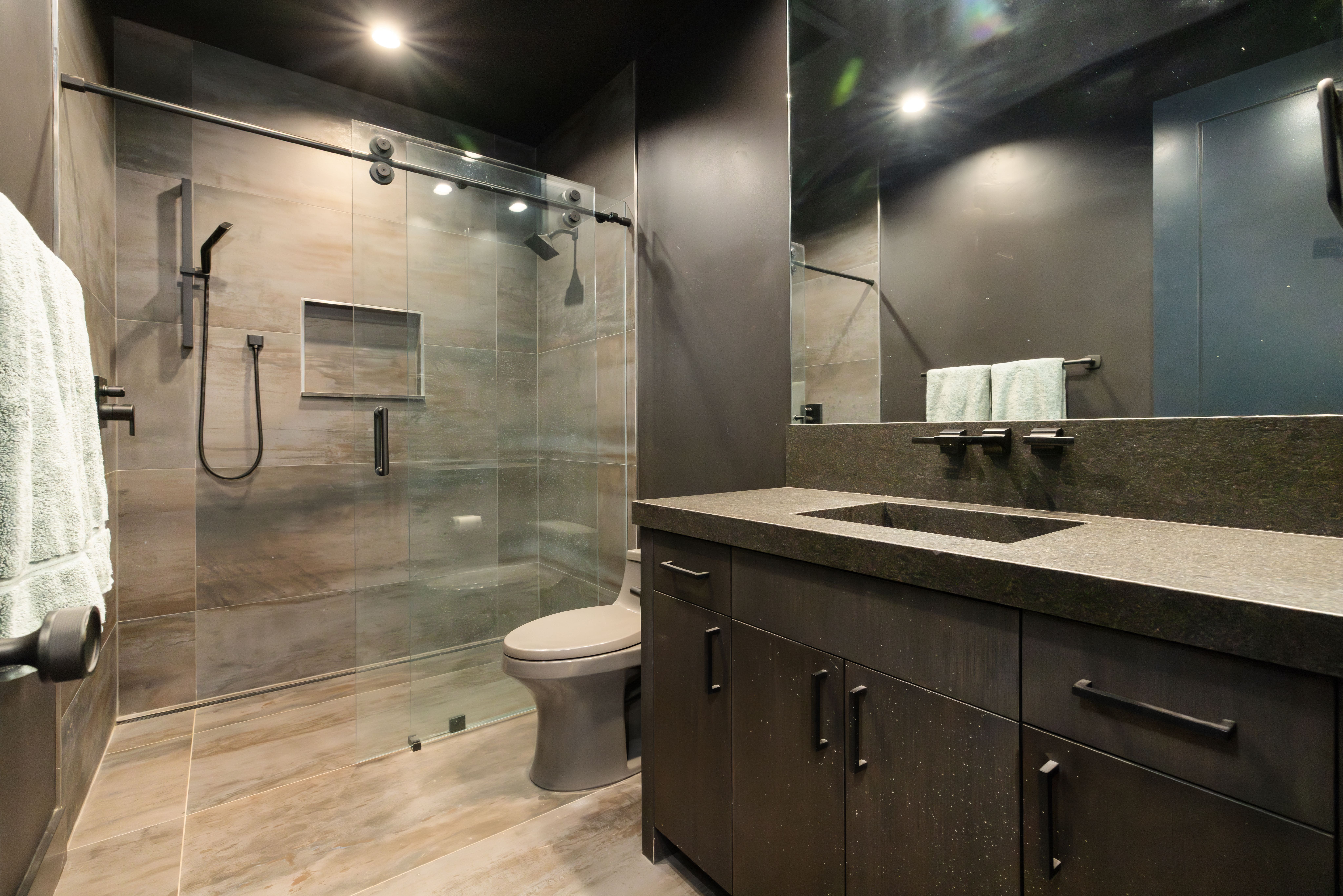 This modern primary bathroom features a bold, monochromatic design with dark, textured cabinetry and a stone-topped vanity. The shower area is enclosed by a clear glass sliding door and includes a wall-mounted niche and an integrated rainfall showerhead. The overall aesthetic is sleek, minimalist, and sophisticated, emphasizing luxury through its dark color palette and clean lines.