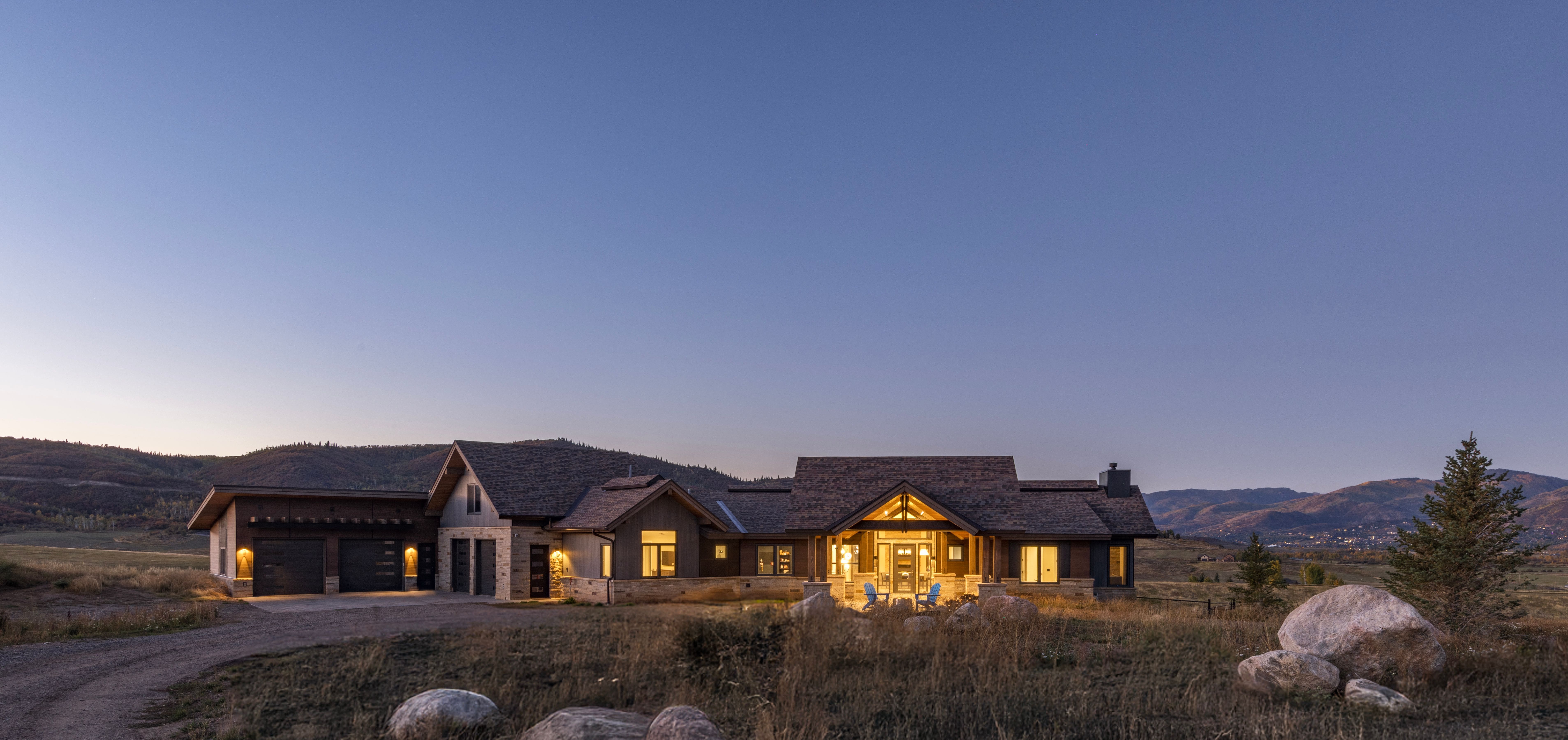 This stunning custom mountain-style home is captured at dusk, showcasing a harmonious blend of stone and dark wood siding architecture. The expansive facade features multiple garage bays and a grand, illuminated entryway, set against a backdrop of rolling hills and a dusky, gradient sky. Manicured yet natural landscaping with large boulders completes the serene, high-end mountain aesthetic.