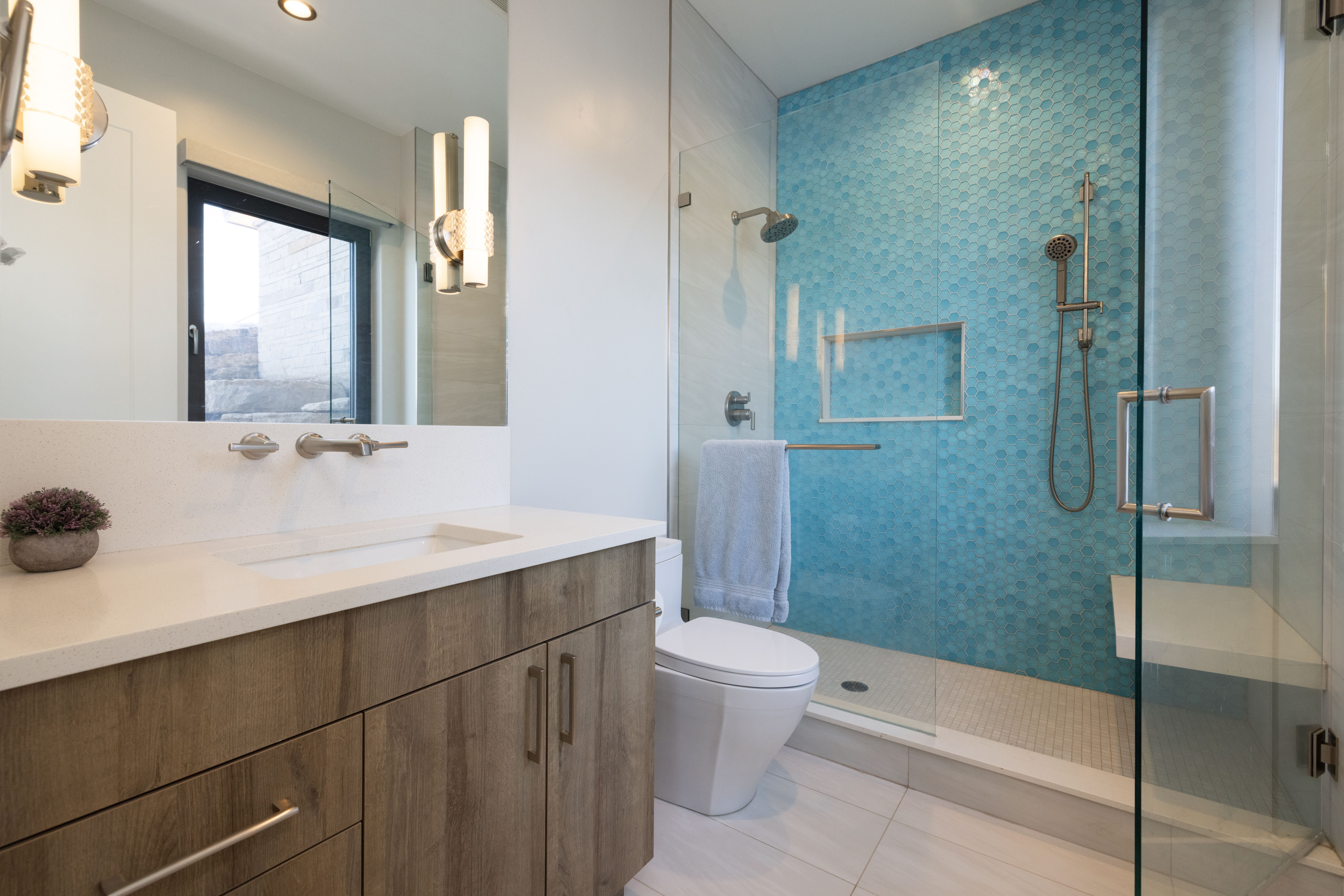 This modern primary bathroom features a wood-vanity with a white quartz countertop and wall-mounted faucets. The walk-in shower is highlighted by striking blue hexagonal tile, a glass enclosure, and a built-in stone bench. A clean, contemporary aesthetic is achieved through high-quality materials and thoughtful lighting, making the space feel both functional and luxurious.