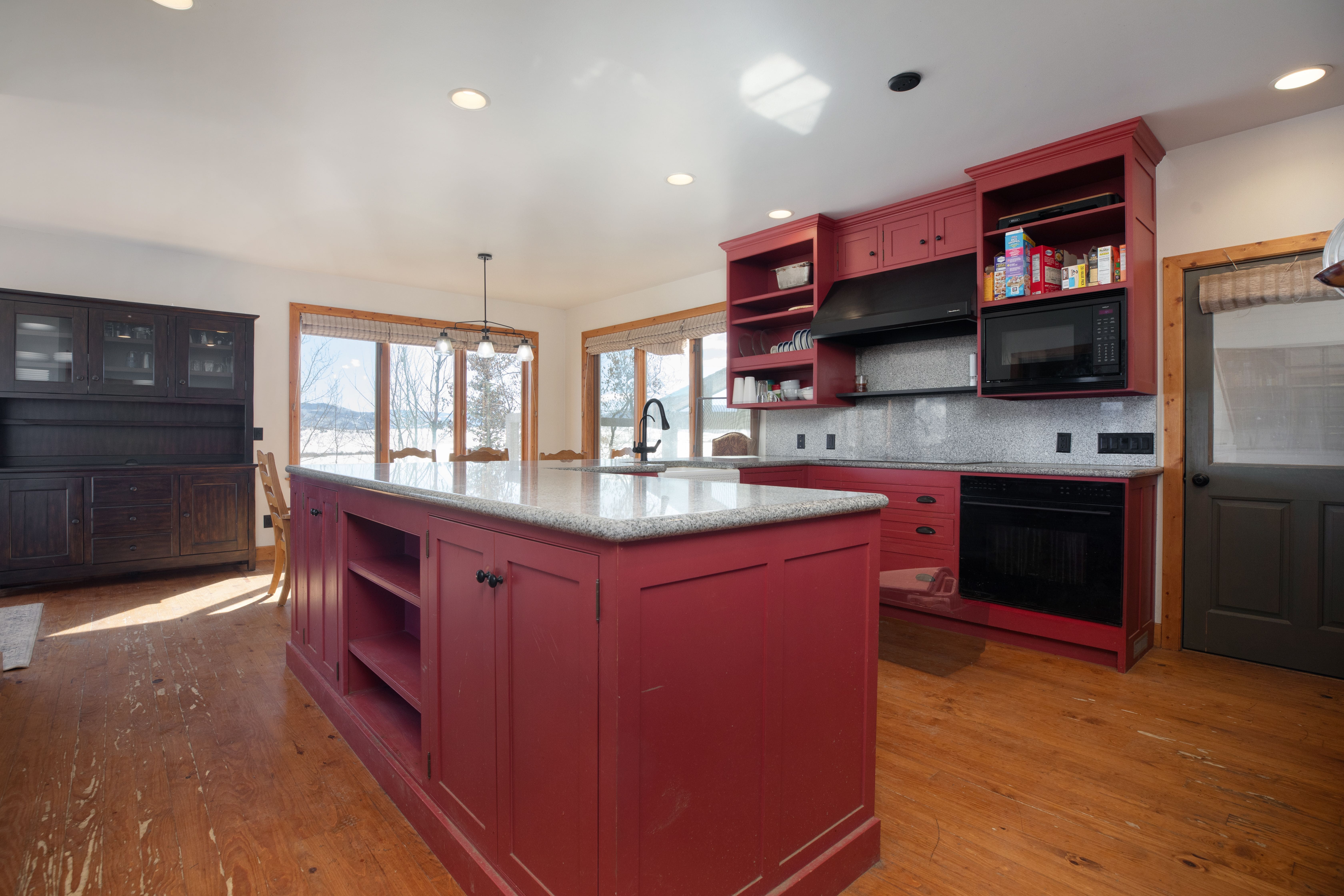 This kitchen features a distinct, rustic style with bold red cabinetry and a spacious central island topped with a speckled granite countertop. The kitchen includes built-in black appliances, open shelving, and a dark wood matching hutch, all set against natural hardwood flooring. Large windows provide ample natural light and a view of the outdoors, creating a cozy and functional cooking space.