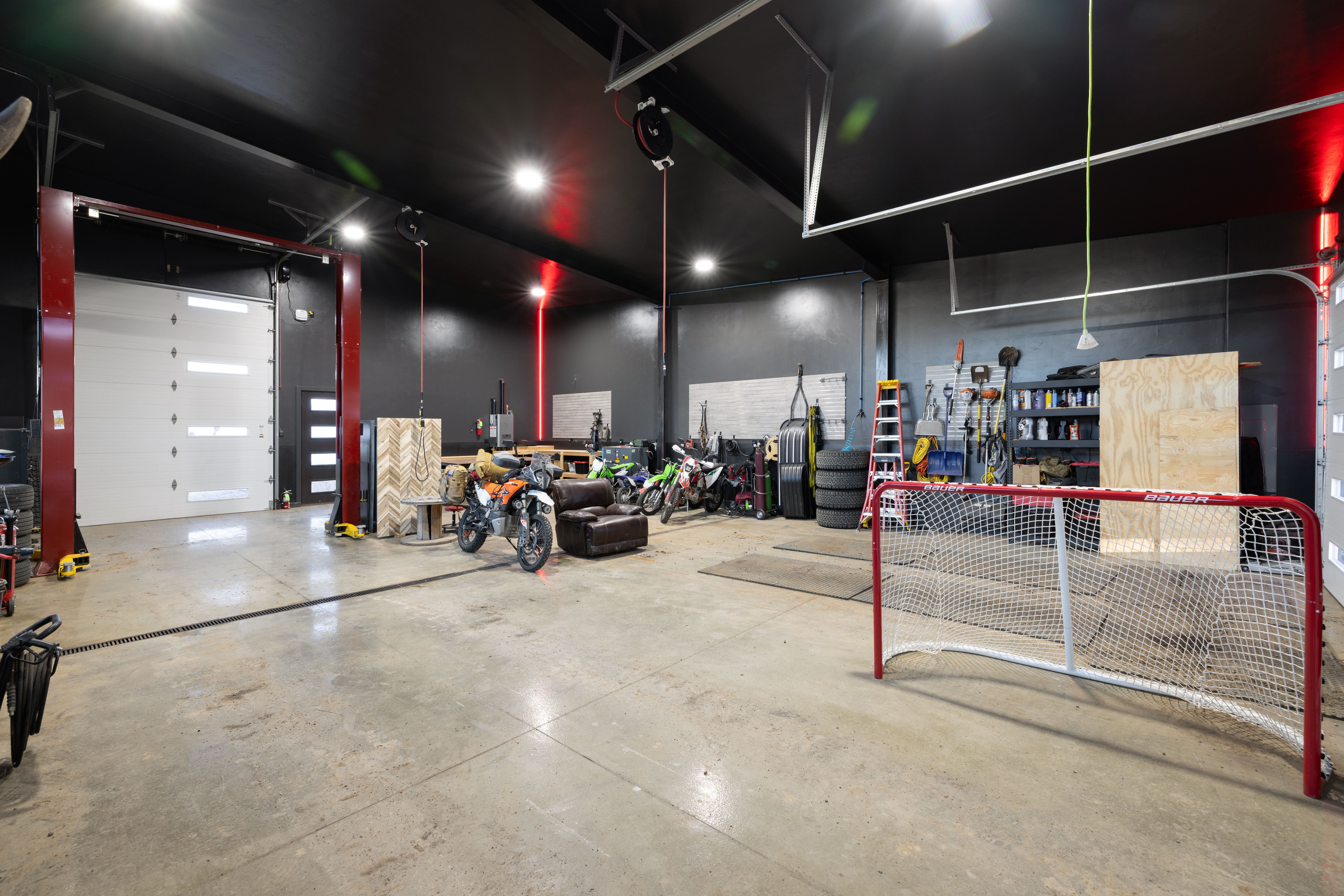 This spacious, high-ceilinged garage features a sleek, modern aesthetic with matte black walls and polished concrete floors. The area is well-outfitted for hobbyists, including a vehicle lift, multiple motorcycles, a storage organization system with tools, and a hockey goal. The industrial design is both functional and versatile, making it an ideal space for automotive work, storage, or recreational activities.