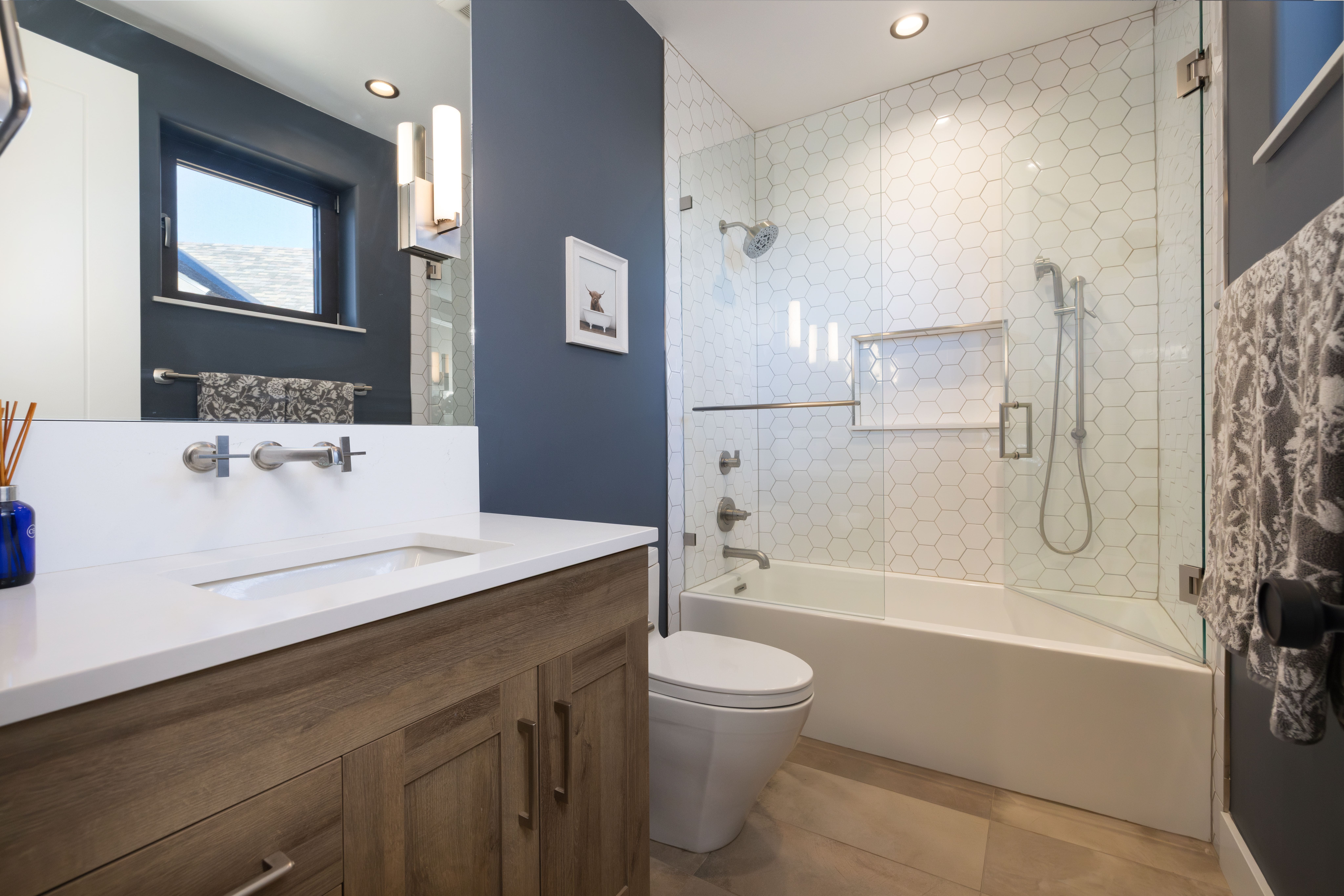 This modern bathroom features a sleek wood vanity with integrated white countertops and a wall-mounted faucet, set against a bold dark blue accent wall. The shower-tub combination is elegantly tiled with white hexagonal tiles and secured by a framed glass enclosure. The space offers a sophisticated, contemporary aesthetic enhanced by warm lighting and high-quality fixtures.