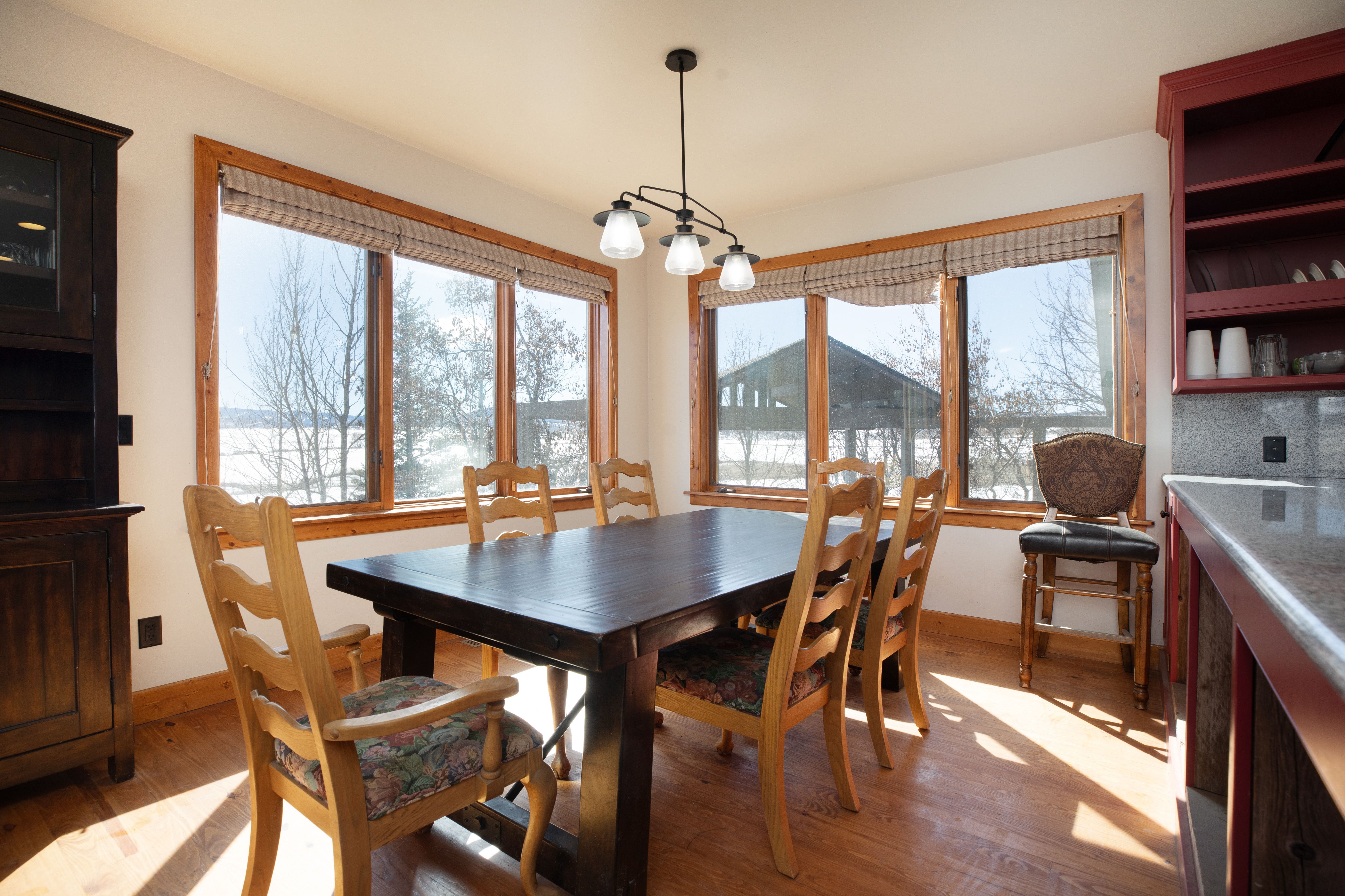 This dining room features a rustic dark wood dining table surrounded by six wooden chairs with floral patterned cushions, set against a backdrop of large windows offering scenic outdoor views. To the left is a tall matching hutch, and to the right, a built-in red cabinet provides added utility and warmth to the space. The room is completed with light wood flooring and a classic three-light overhead chandelier, creating a cozy and inviting atmosphere.