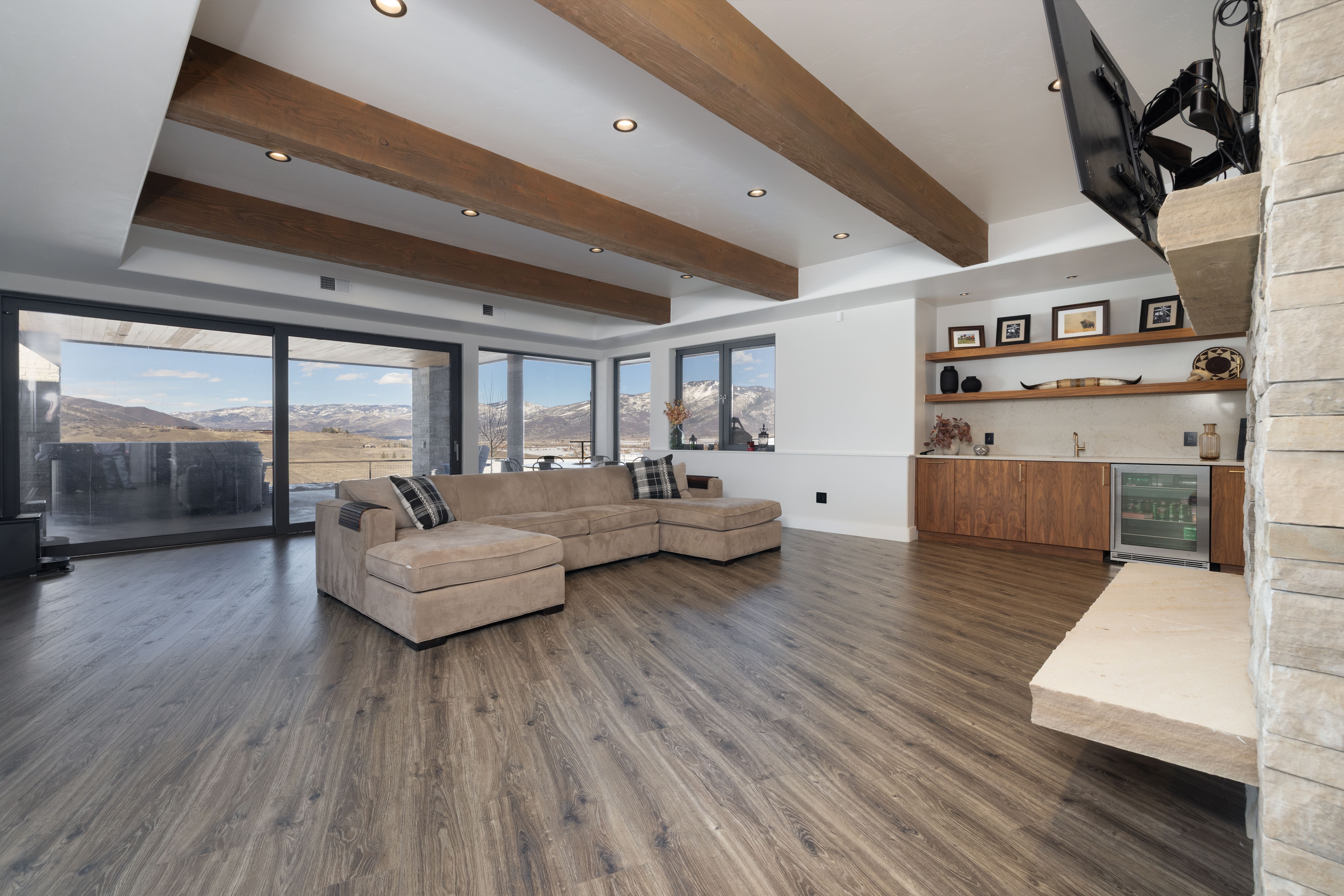 This spacious, open-concept living area features warm wood-toned flooring and eye-catching exposed wood beams across the ceiling. The room is centered around a large, comfortable L-shaped sectional sofa and boasts a built-in wet bar area with stone shelving and an under-counter beverage fridge. Expansive sliding glass doors provide seamless indoor-outdoor living, framing stunning, panoramic mountain views.