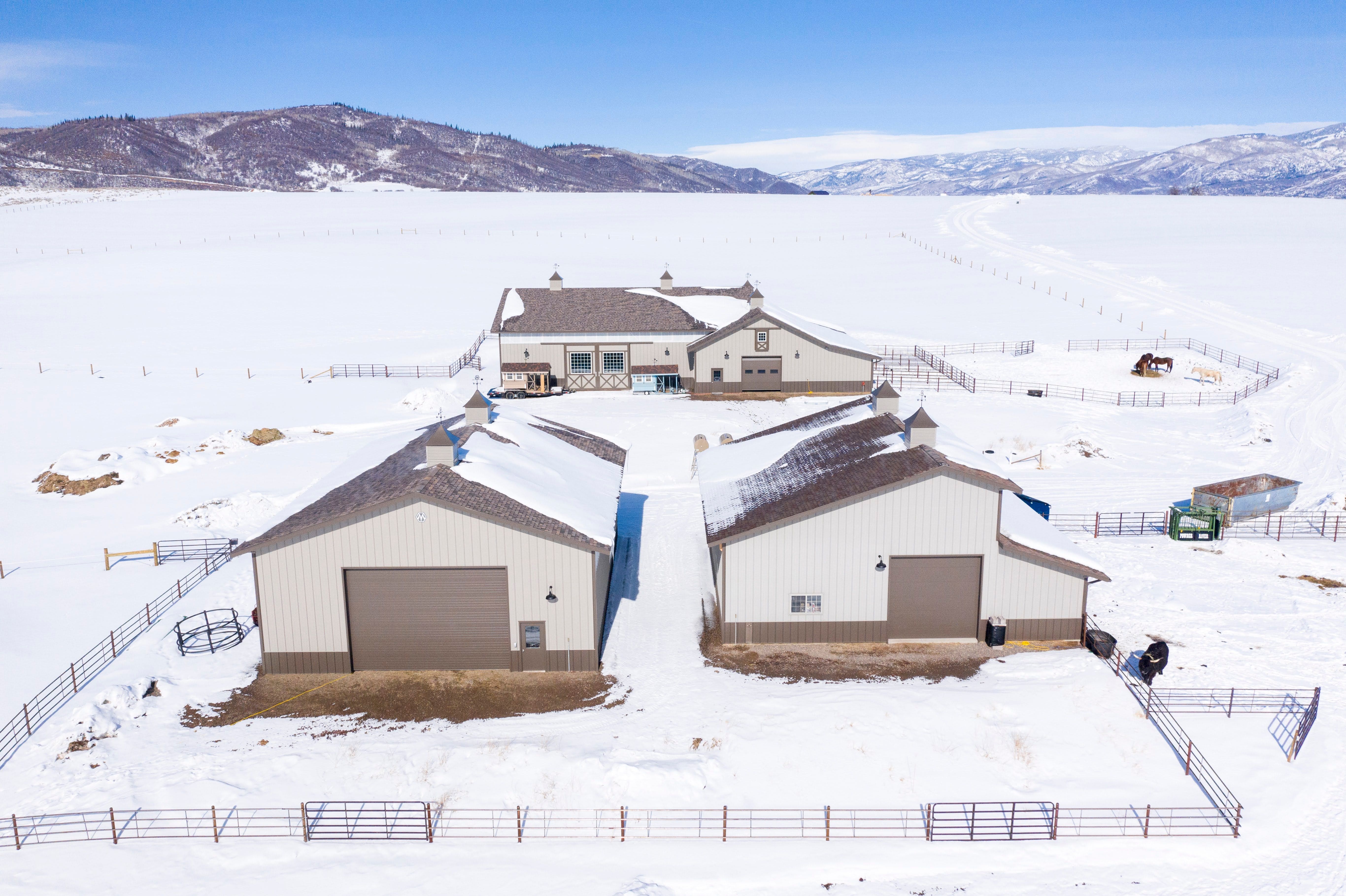 This aerial view showcases a rural property highlighted by three matching light-colored outbuildings with dark brown roofs, situated on a sprawling, snow-covered landscape. The property is well-defined by extensive metal fencing, creating separate enclosures for livestock, while rugged mountains flank the horizon in the background. It is a functional and well-maintained equestrian or ranch-style facility perfect for those seeking space and utility in a cold-climate environment.